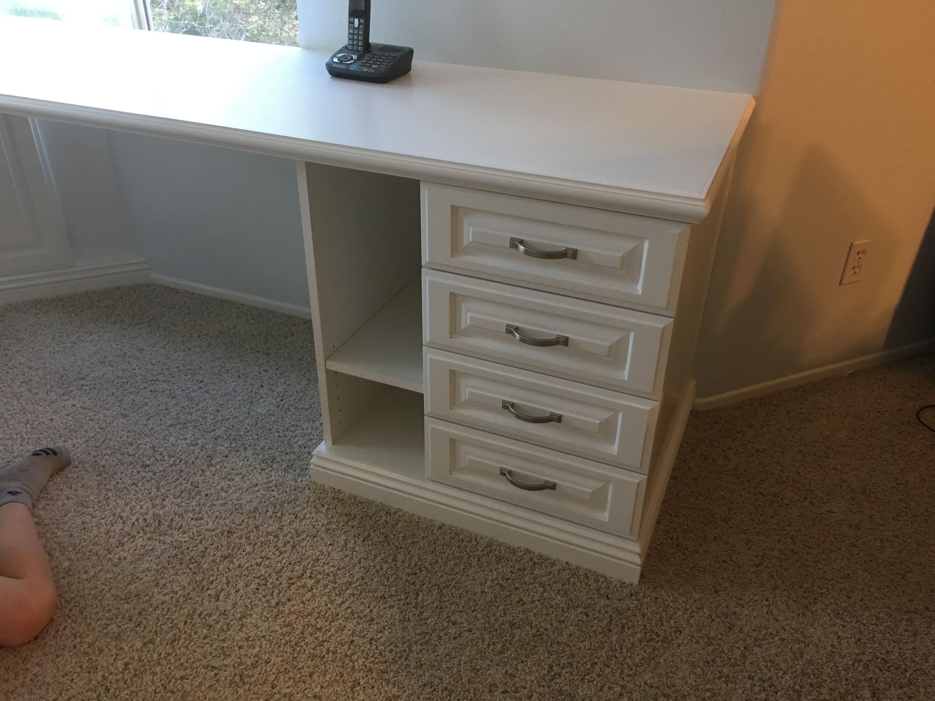 White desk with drawers and open shelf in a room with tan carpet.