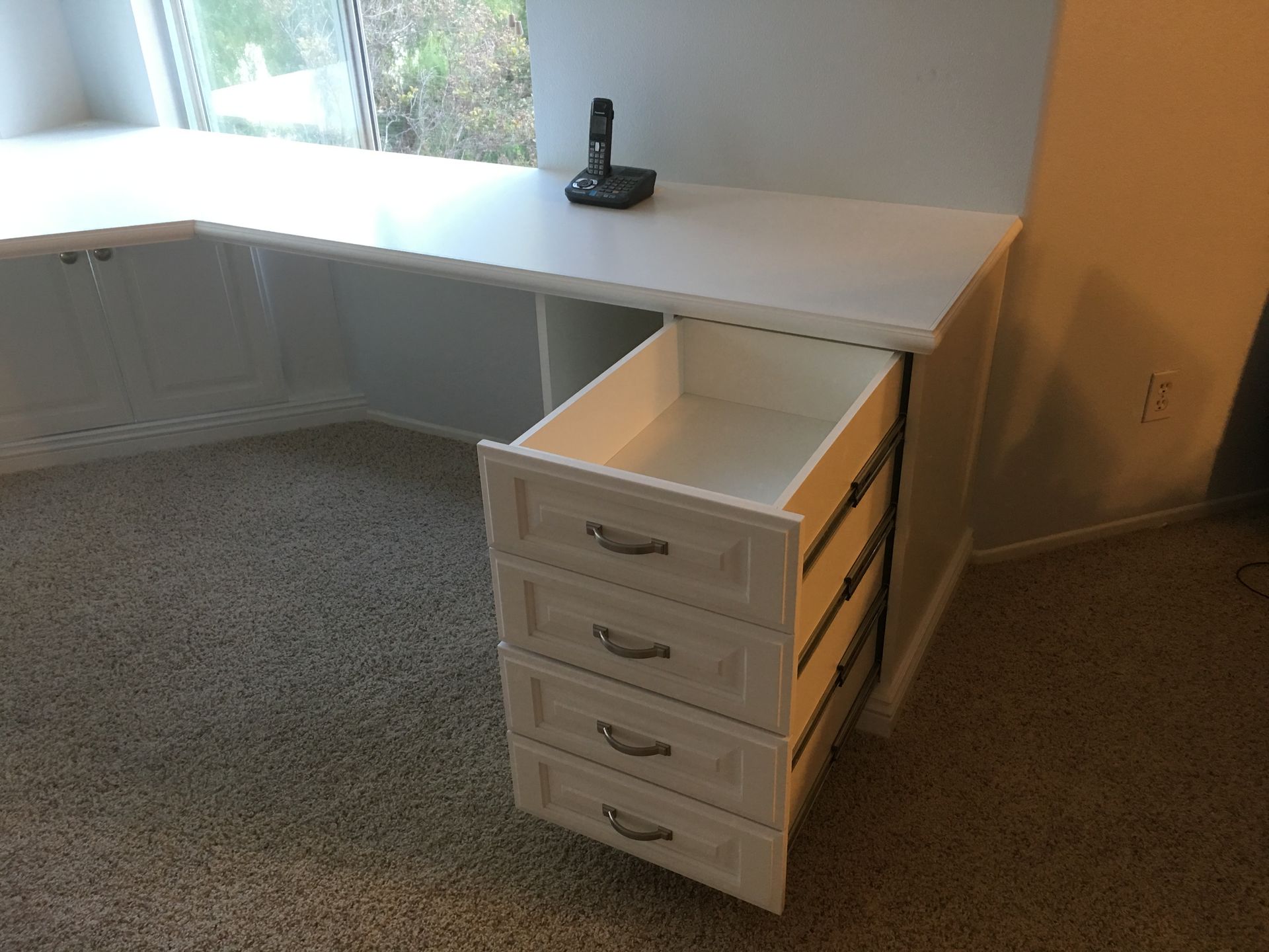 White desk with open drawer, telephone on top. Room with carpet, next to a window seat.
