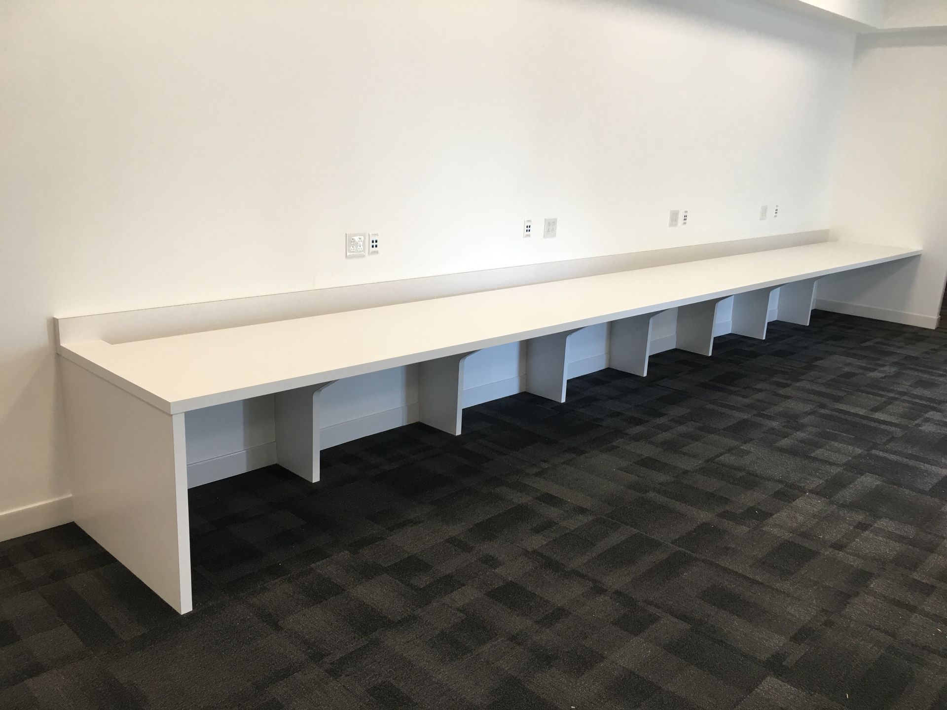 Long white desk against a white wall with individual cubby spaces, in a room with patterned carpet.