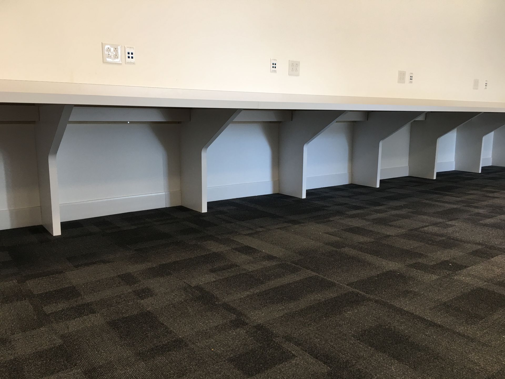 White desks with supports along a wall with power outlets; dark gray patterned carpet.