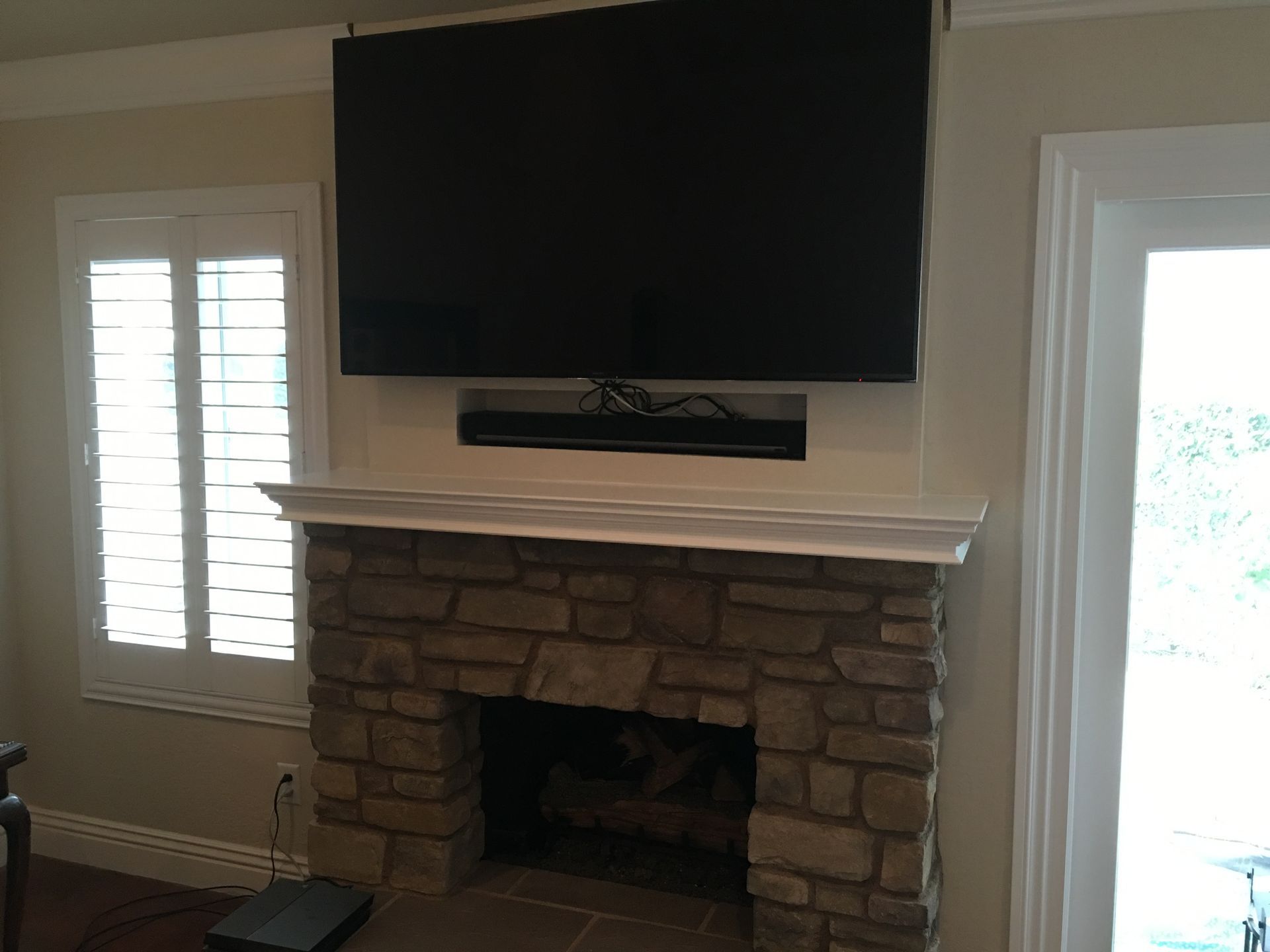 A mounted TV above a stone fireplace with a soundbar. Shuttered window to the left.