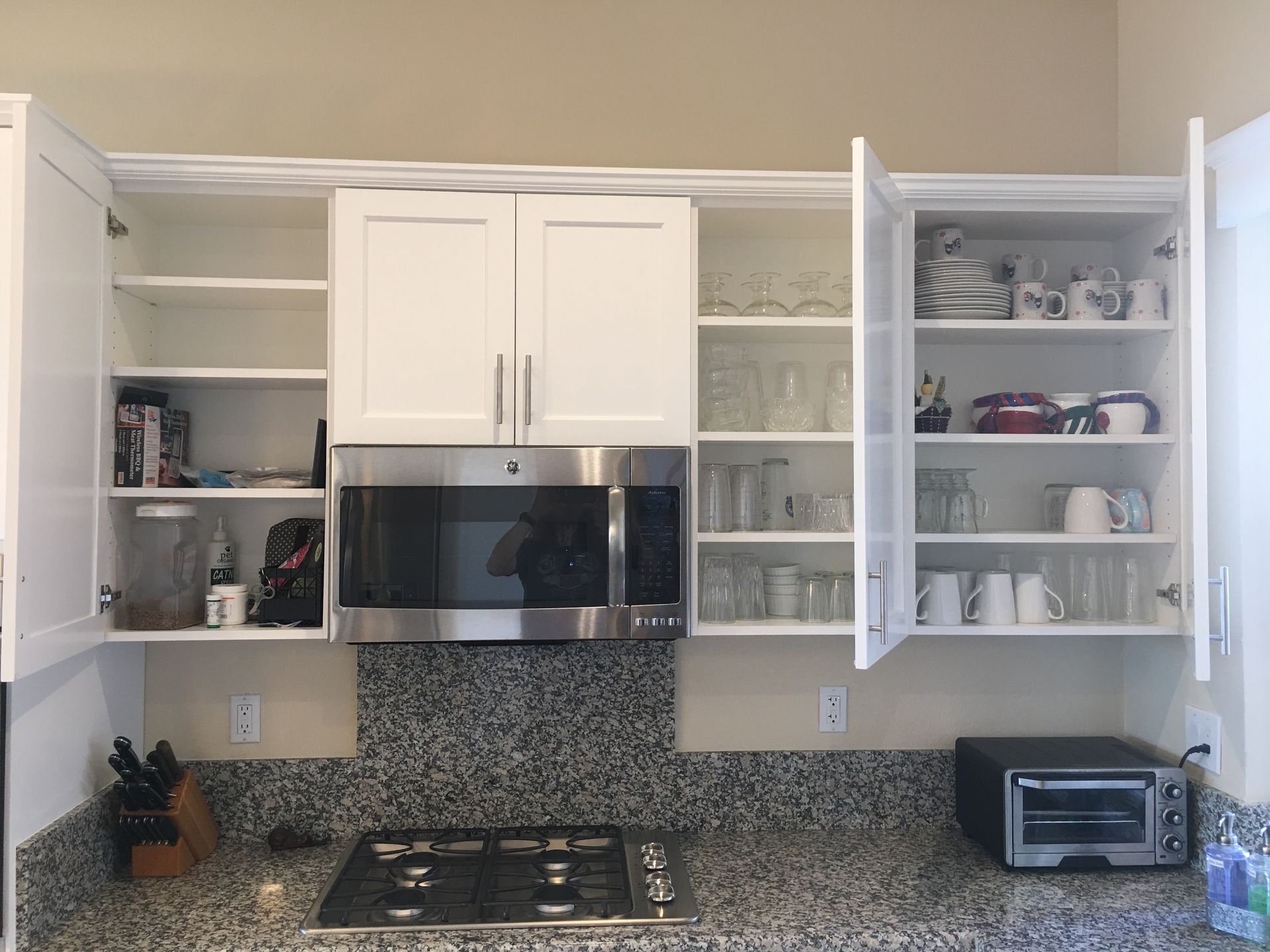 White kitchen cabinets open above a microwave and stovetop. Cups and glasses are displayed.