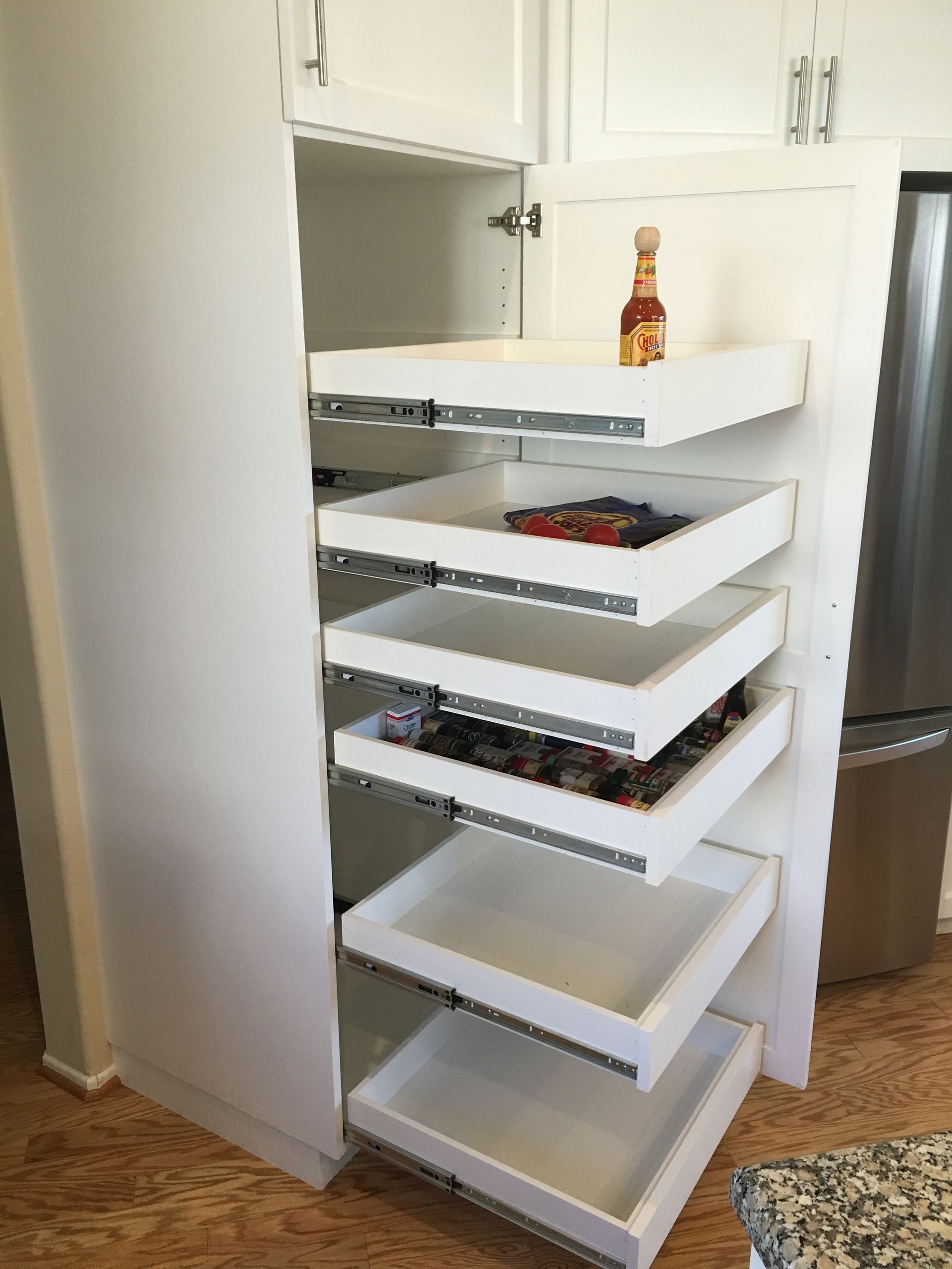 White kitchen pantry cabinet with pull-out shelves, holding food items; hot sauce bottle sits on top shelf.