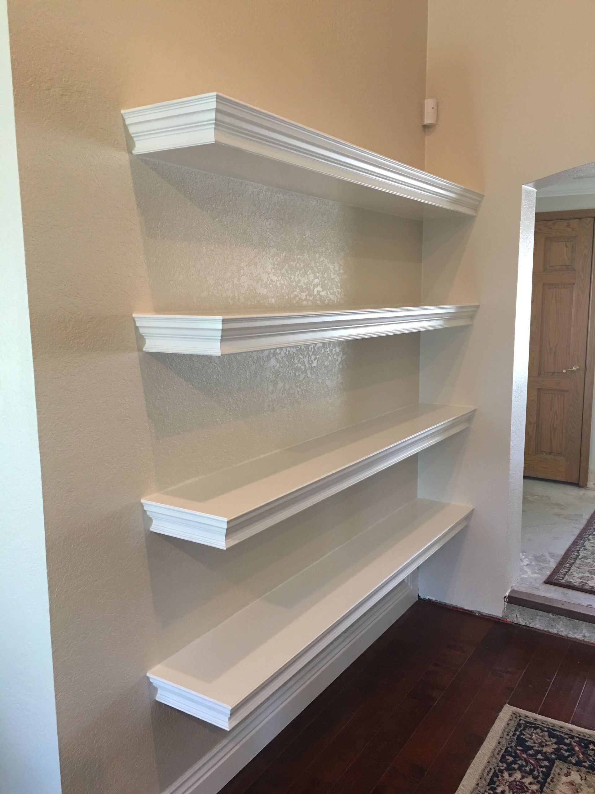 White floating shelves on a beige wall in a room with hardwood floors and a doorway.