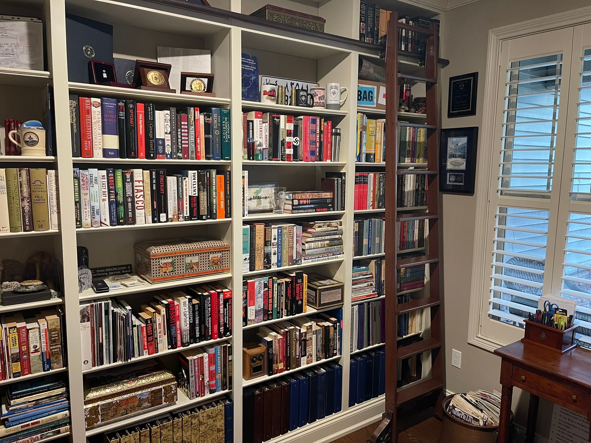 Bookshelves filled with books, a rolling ladder, and a small desk by a window.