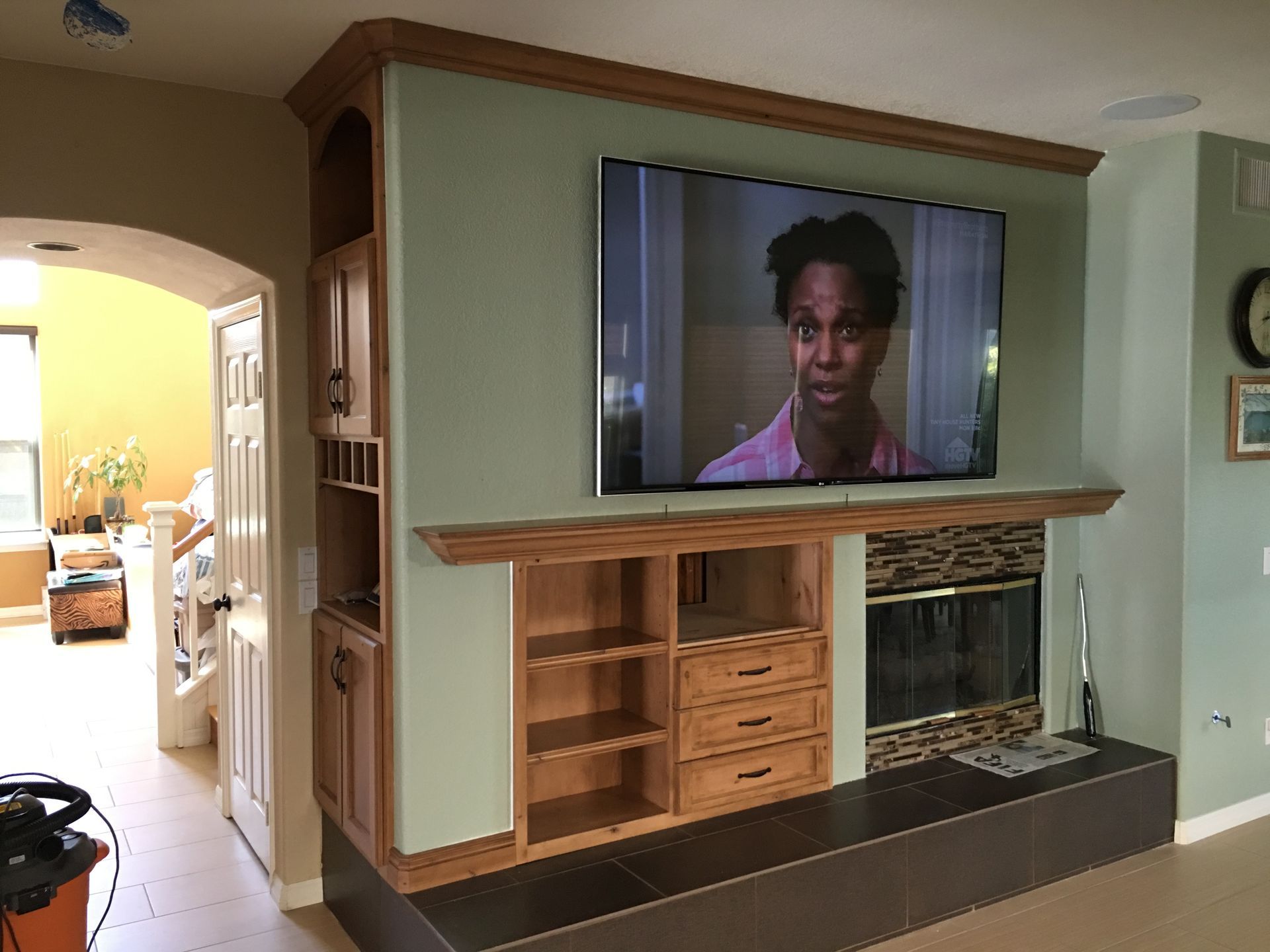 TV mounted above fireplace with built-in cabinetry. Woman on screen, green wall, tan trim.