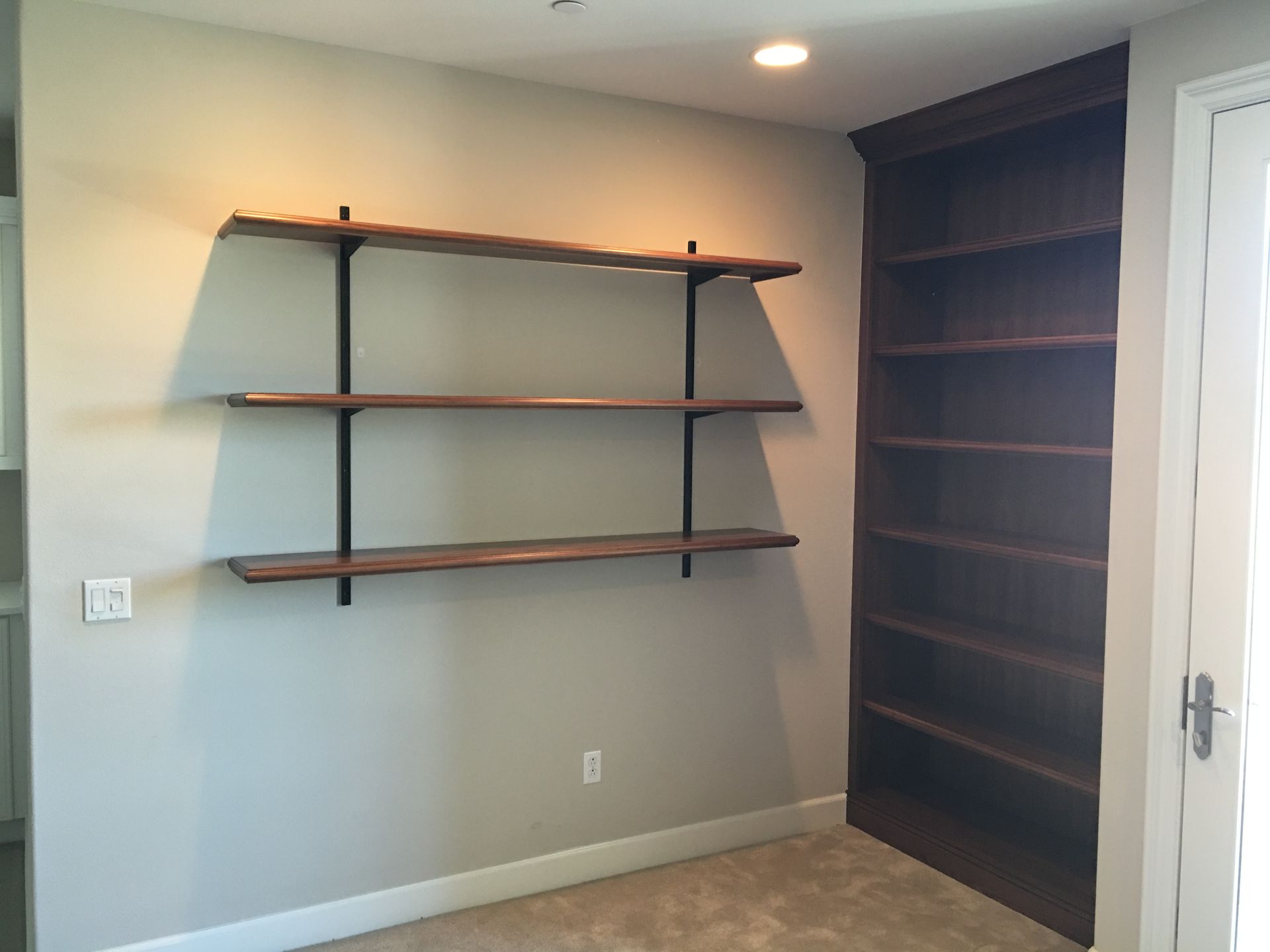 Three wooden shelves with black brackets on a light gray wall, next to a tall, dark wood bookshelf.