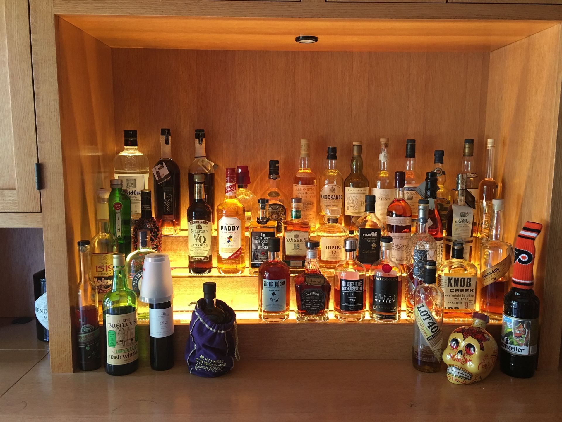 Liquor bottles displayed on tiered shelves in a lit, wooden bar cabinet.