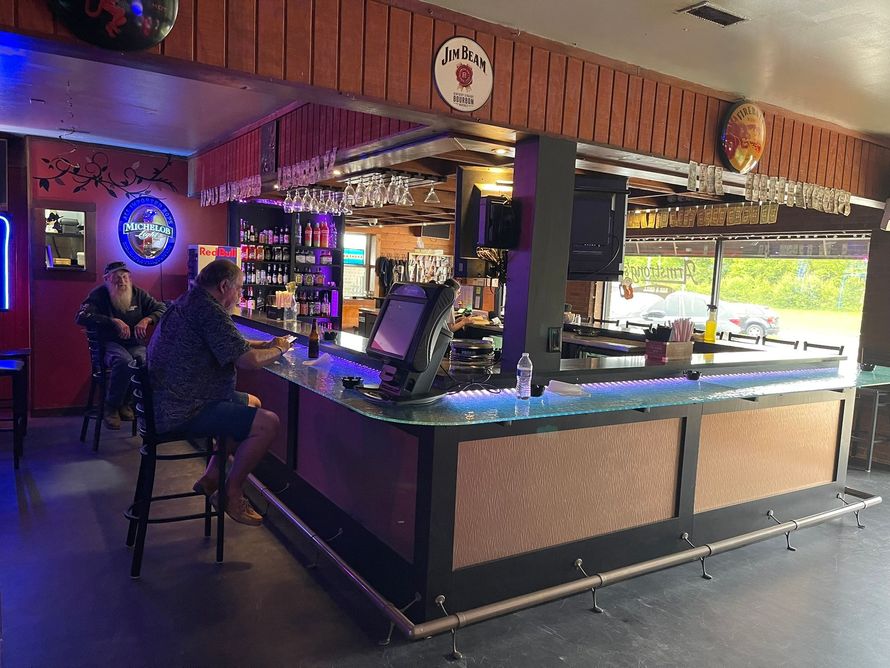 A wooden bar in a dimly lit room, with bottles and a video game visible behind the counter.