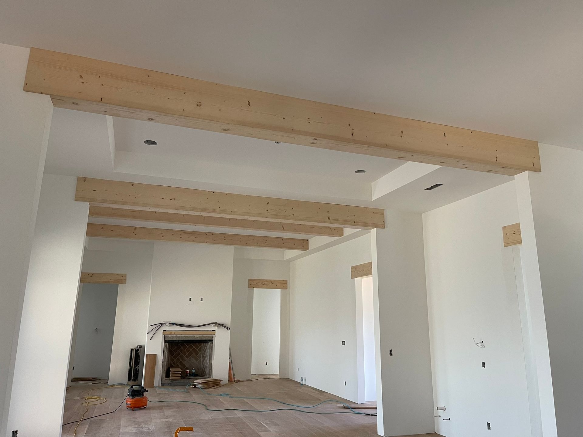 Interior view of a room under construction, featuring wooden beams on the ceiling and doorways. White walls, wooden flooring.