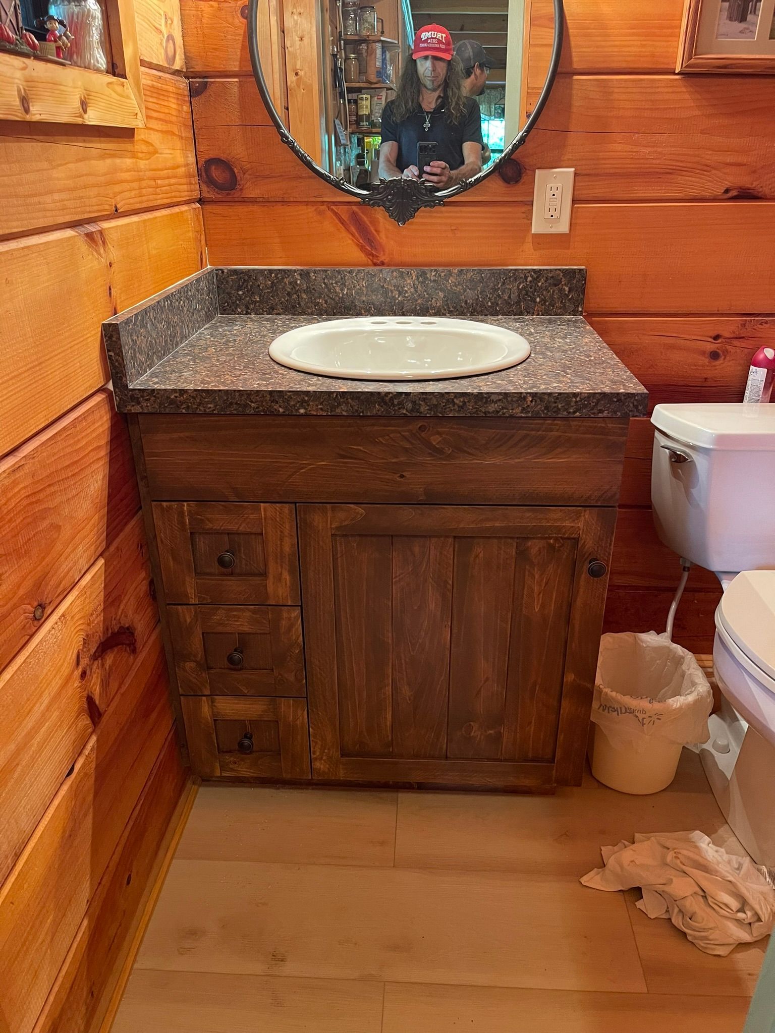 Bathroom with rustic wood vanity, oval sink, dark countertop, and mirror. White toilet and wastebasket are visible.