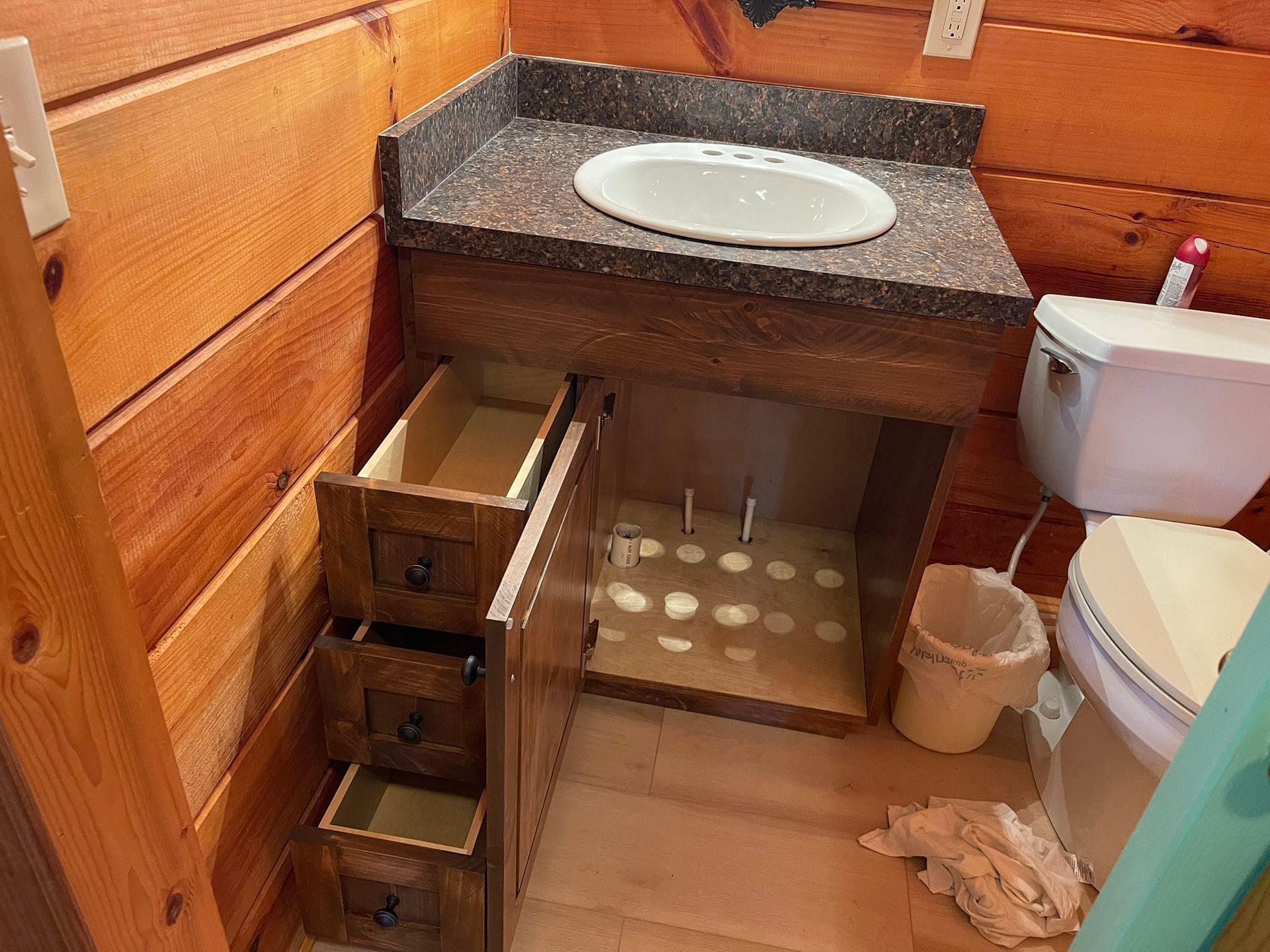Bathroom vanity with open drawers, sink, and toilet in a wood-paneled room.