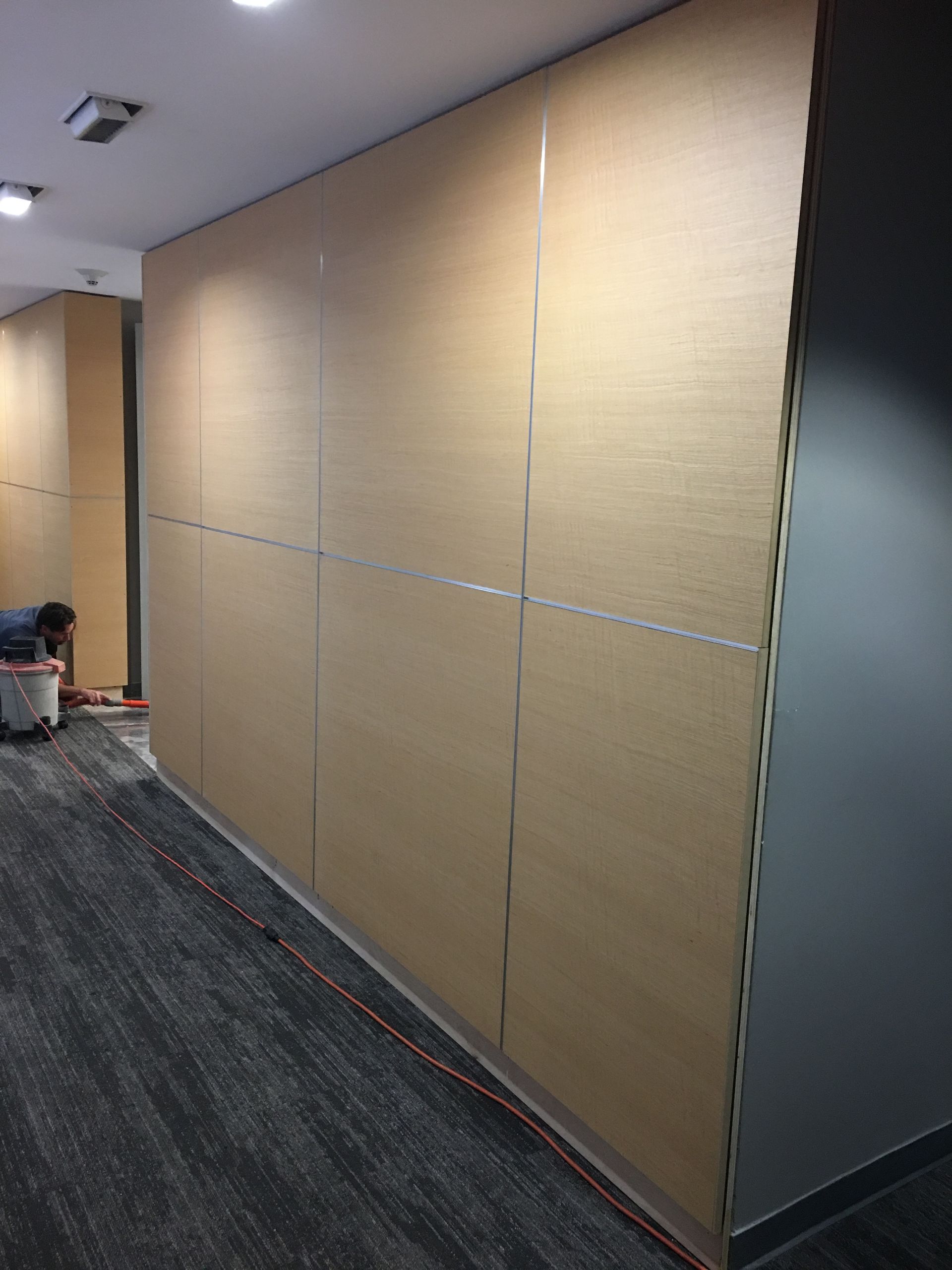 Corridor with light brown paneled wall; worker near unfinished construction, grey carpet, gray wall.