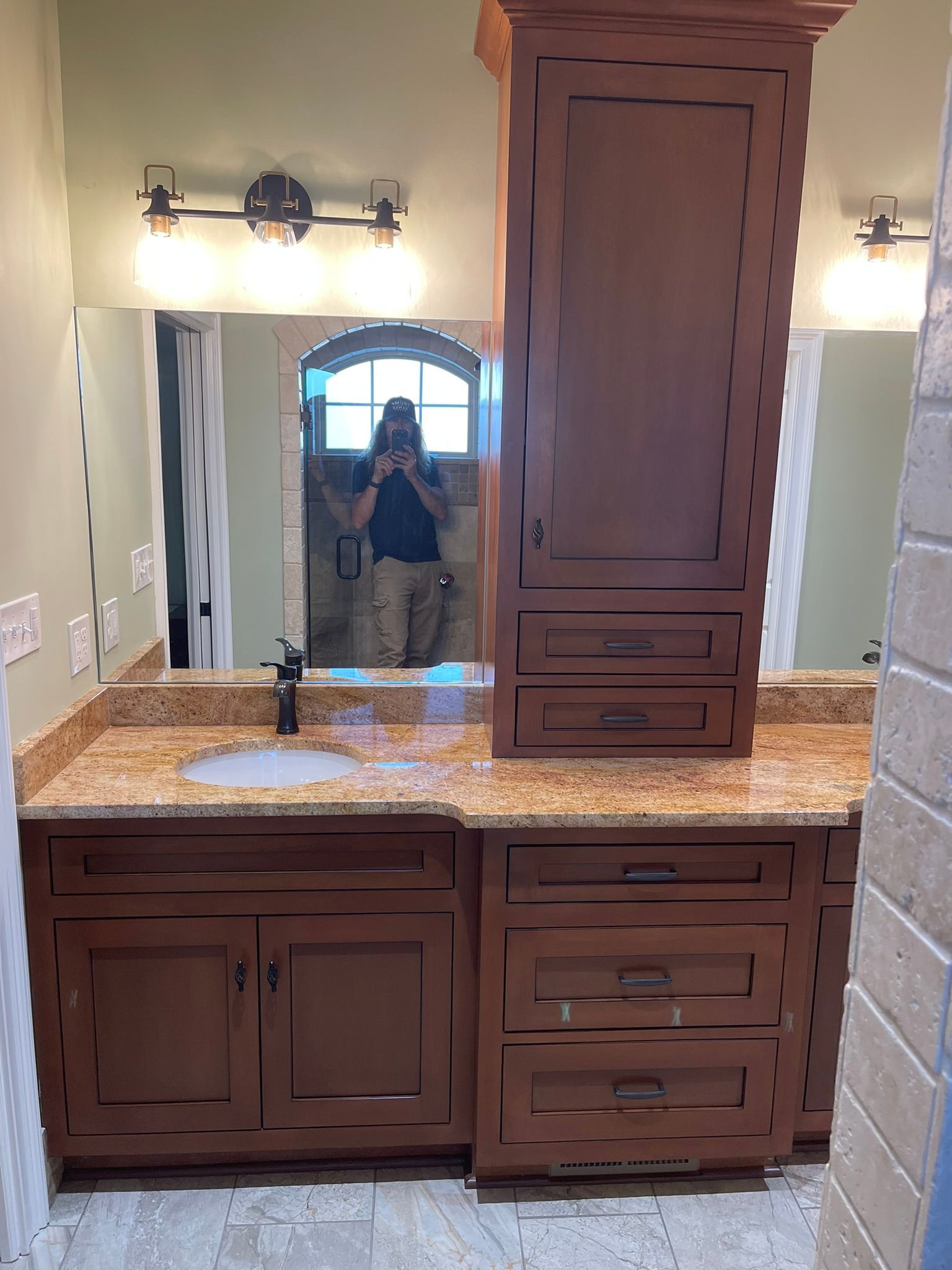 Bathroom with wooden vanity, granite countertop, tall cabinet, and a person reflected in the mirror.