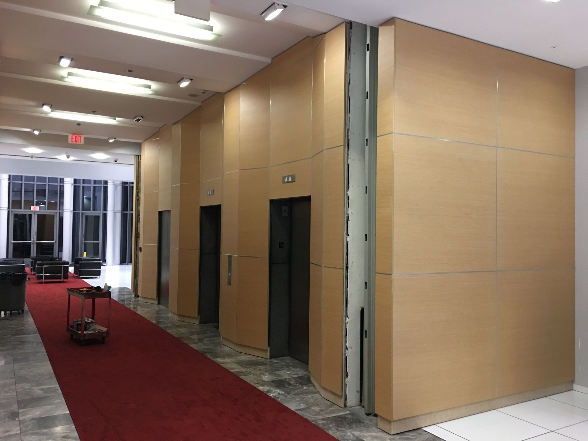 Hallway with three elevators, paneled walls, red carpet, and gray tiled floor.