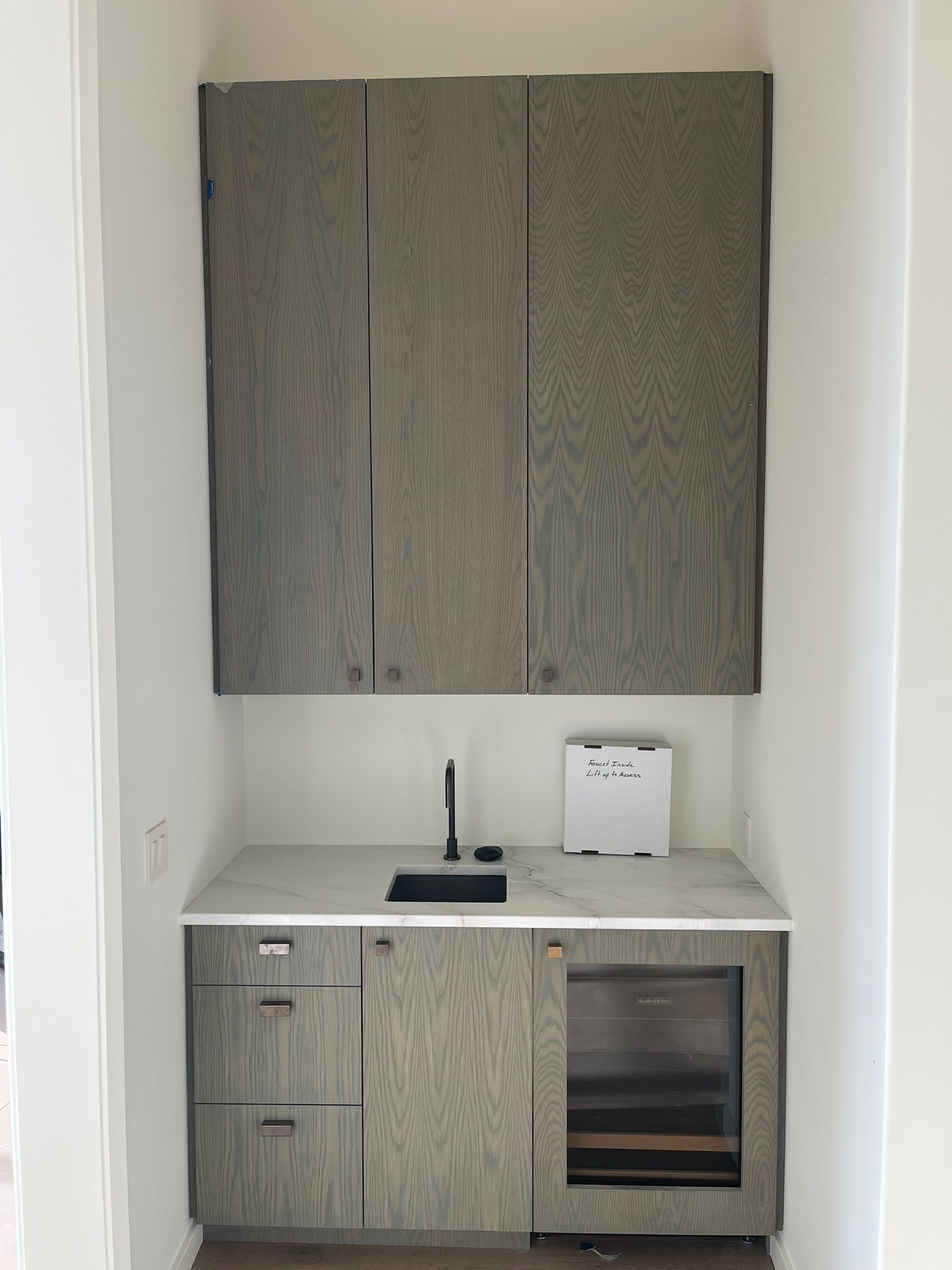 Small built-in bar area with wood grain cabinets, marble counter, sink, and mini fridge in a white alcove.