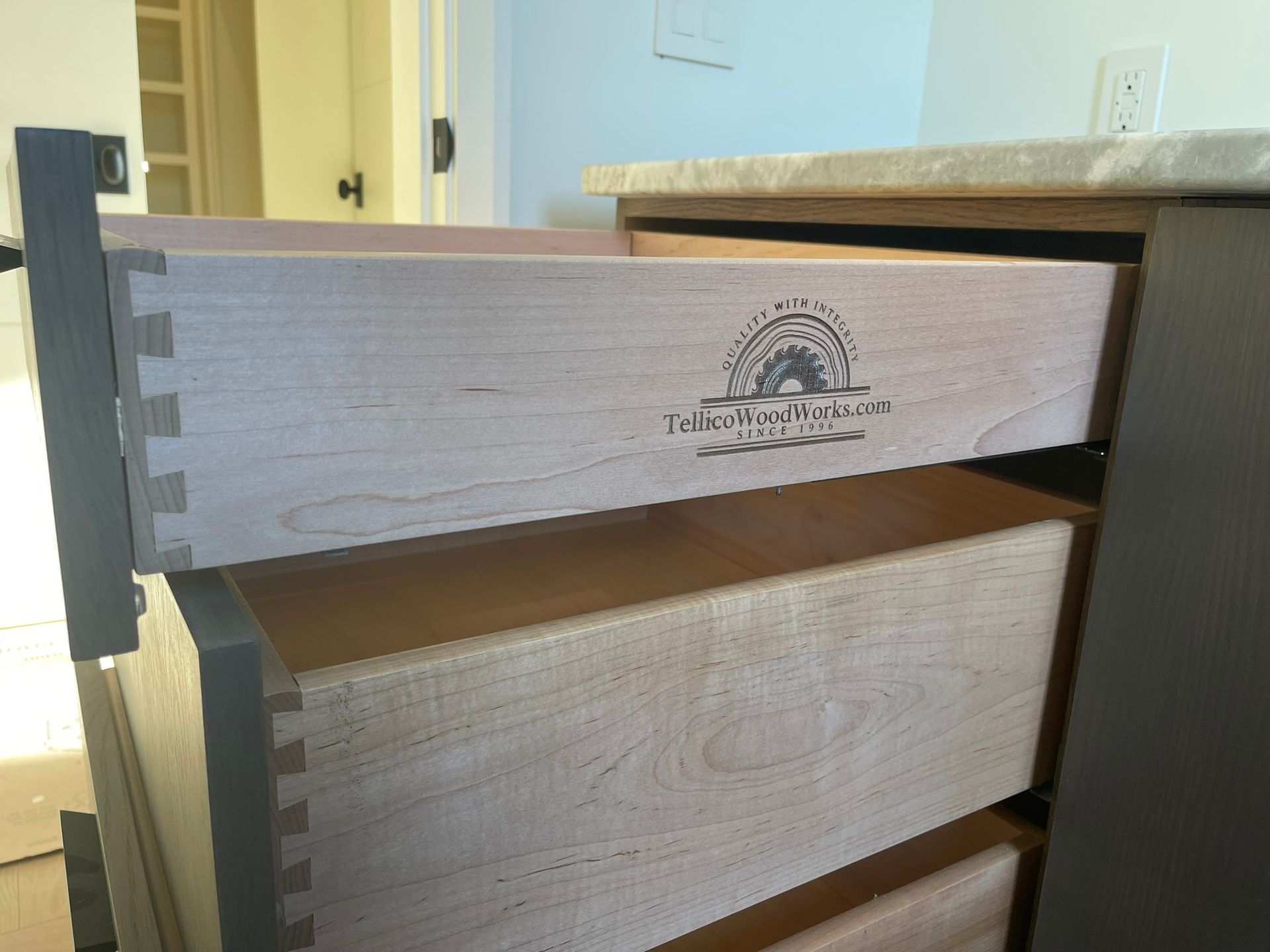 Open wooden cabinet drawer with dovetail joints, stamped logo, and gray hardware.
