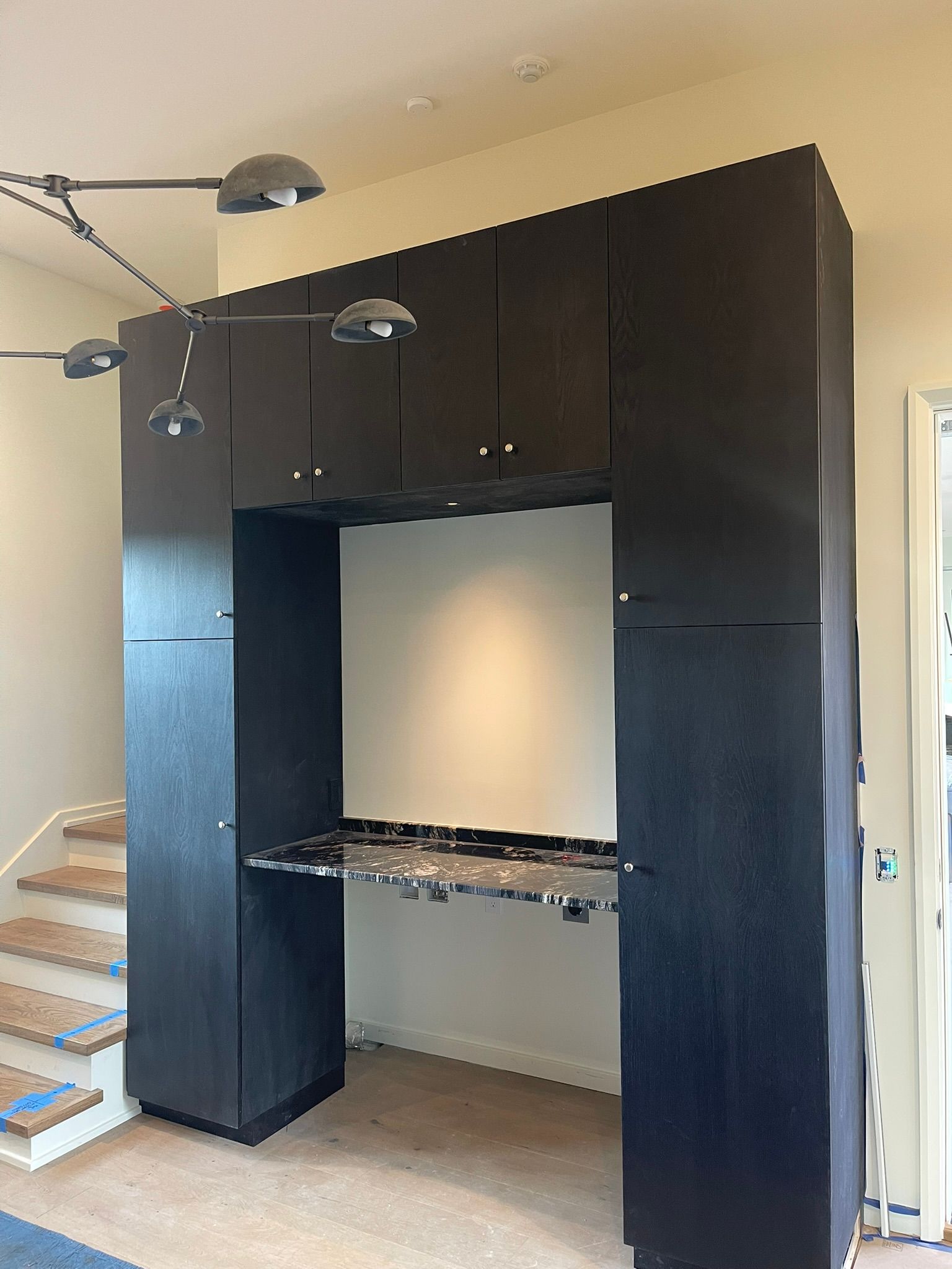 Black built-in desk with upper cabinets, against a white wall. Staircase to the left, pendant lights above.