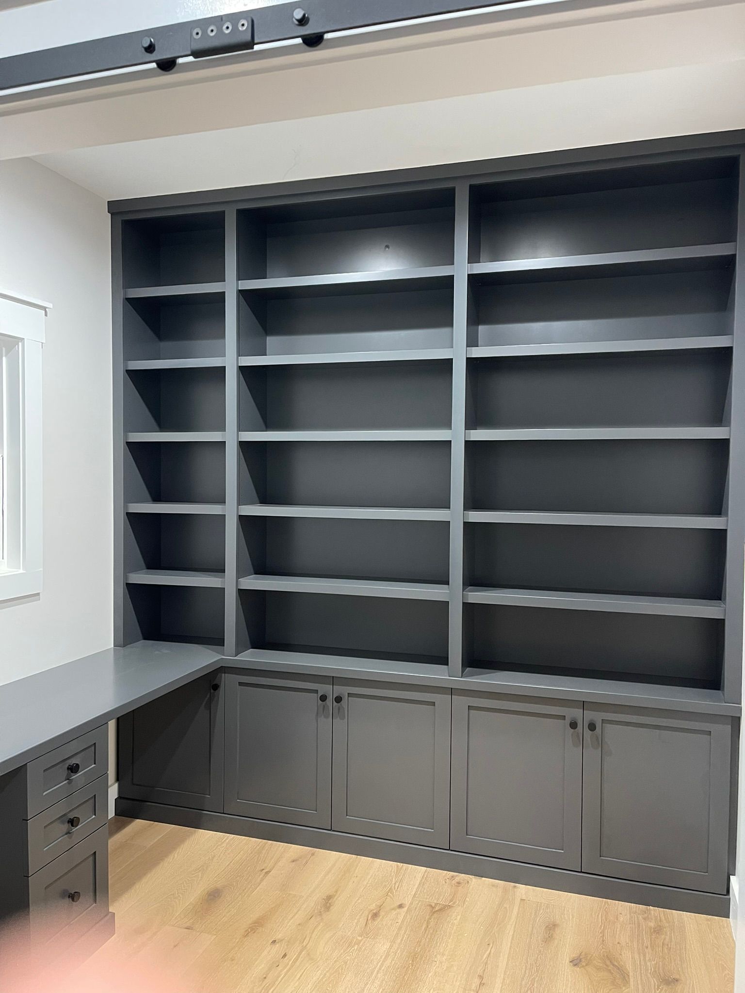 Built-in gray bookcase with open shelves and lower cabinets, attached to a gray desk, in a room.