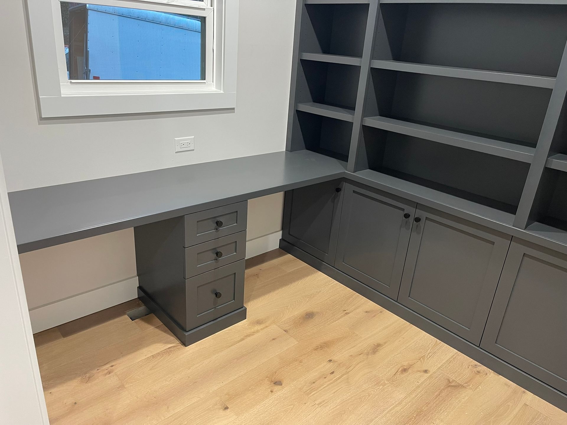 Built-in gray desk and bookshelves in a corner of a room, with a window and wood-look flooring.