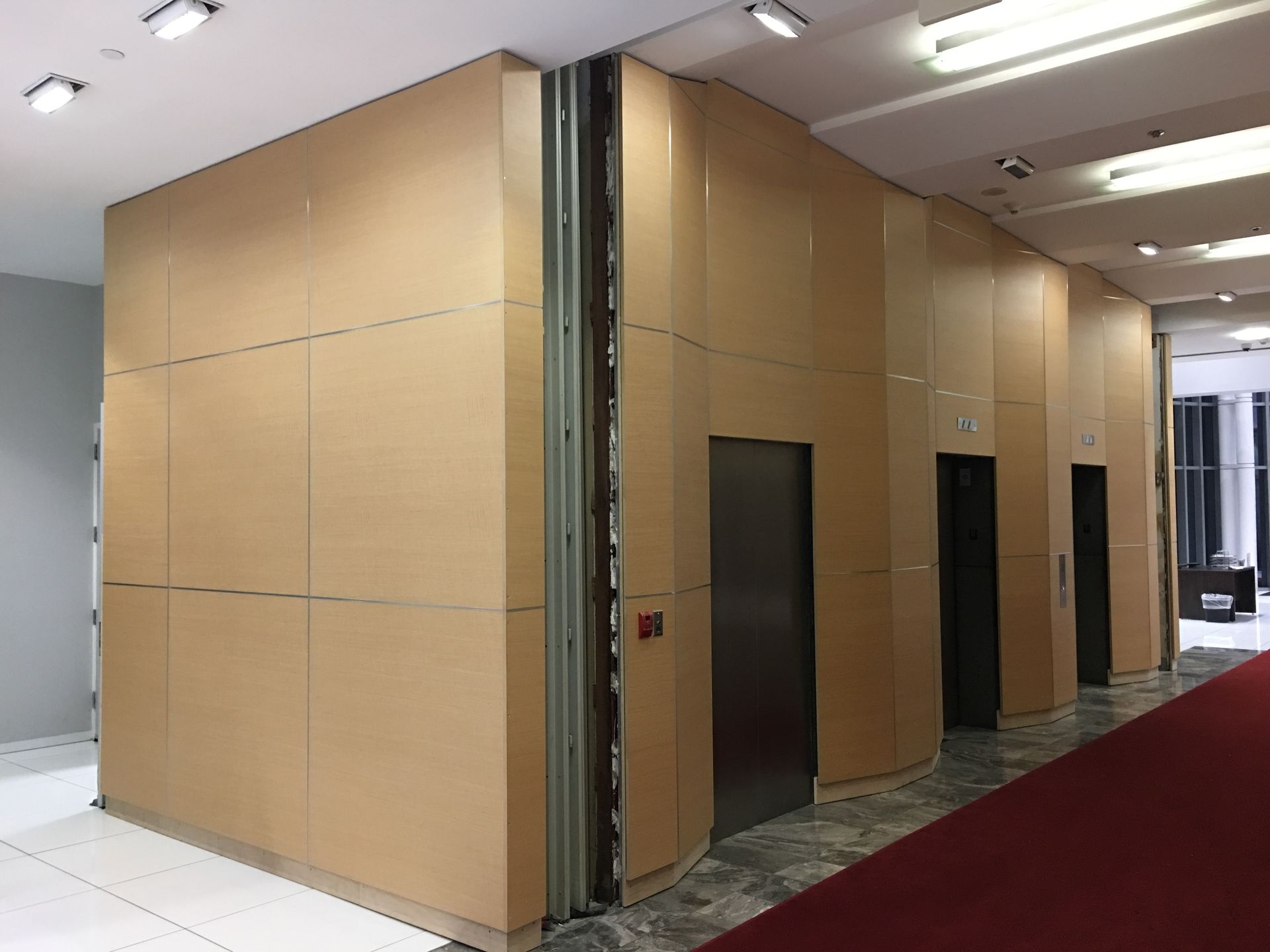 Elevator bank with light tan wood paneling. Doors are stainless steel. Red carpeted floor.