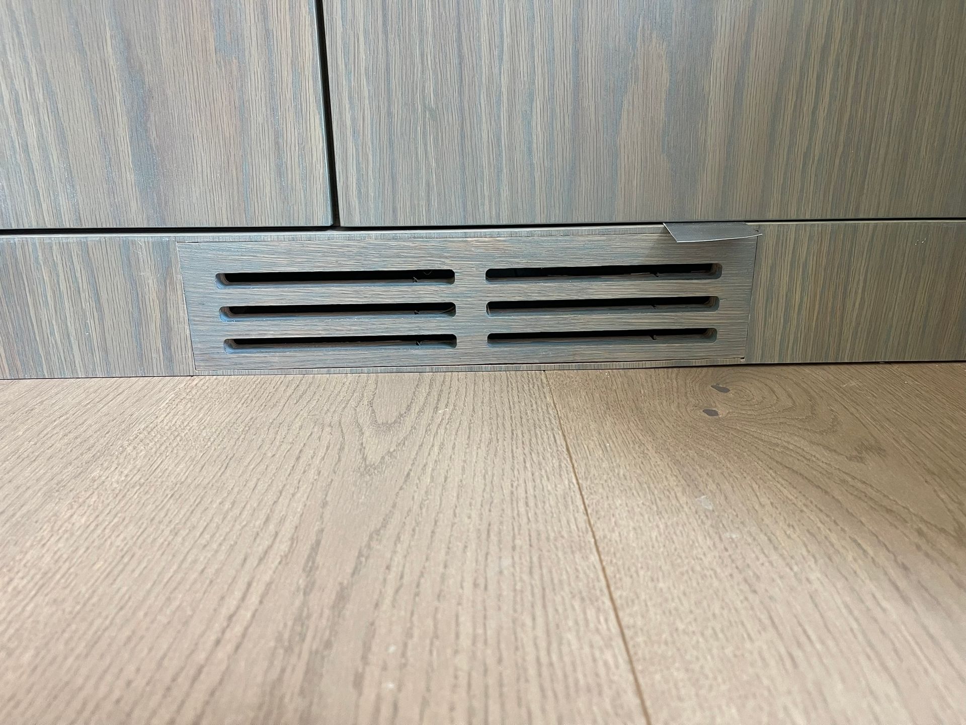 Ventilation grille in wood cabinet base on a wood floor.