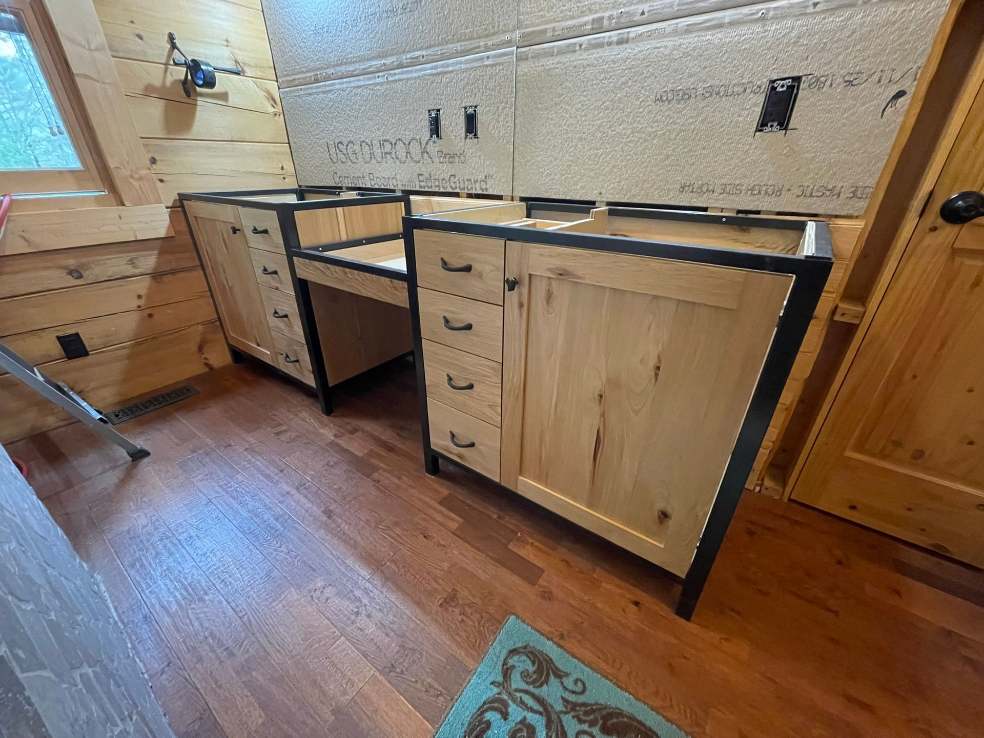 Bathroom cabinets, unfinished wood with black trim, against a wall. Wooden floor, door, and a rug.