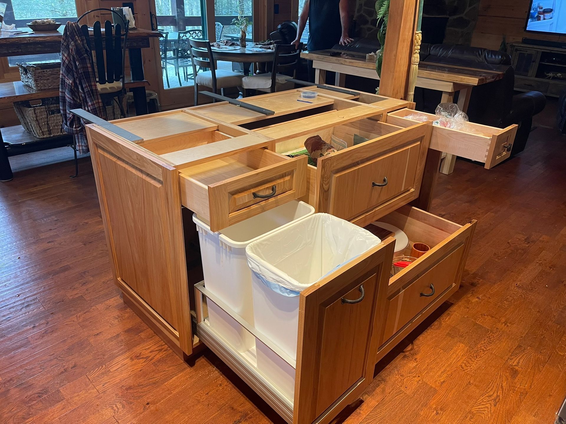 Wooden kitchen island with open drawers and pull-out trash bins on a wood floor.