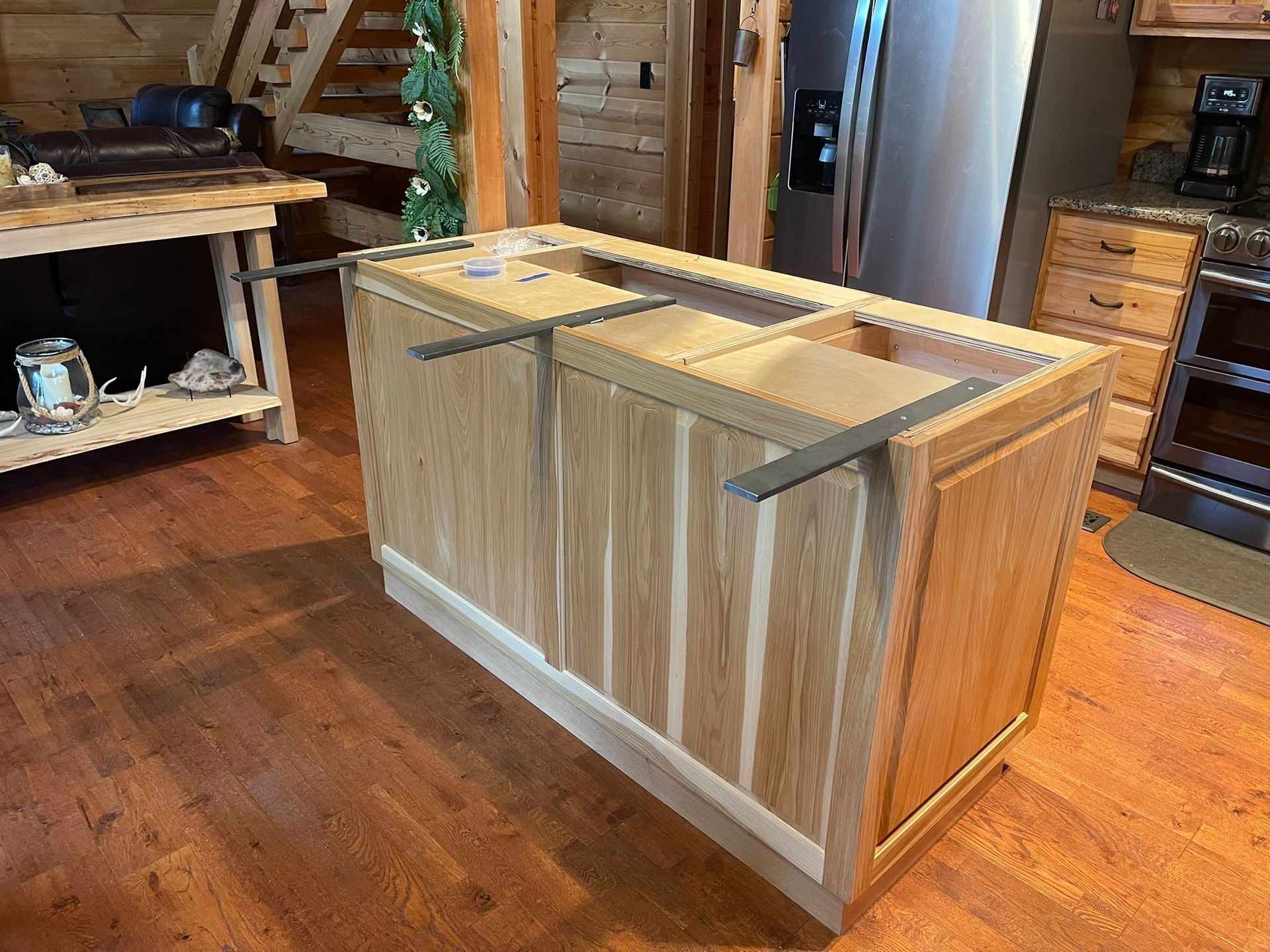 Kitchen island cabinet, light wood, unfinished with metal supports; wood floor and visible kitchen.