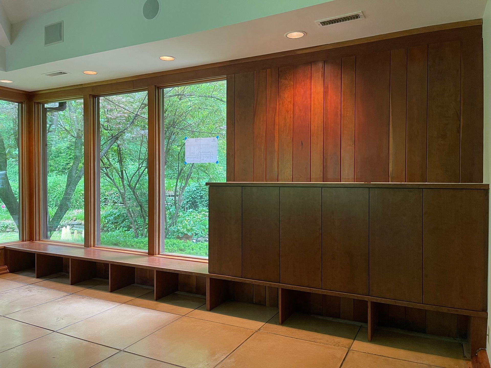 Room with large windows, wooden paneling, and built-in storage. Sunlight streams in.