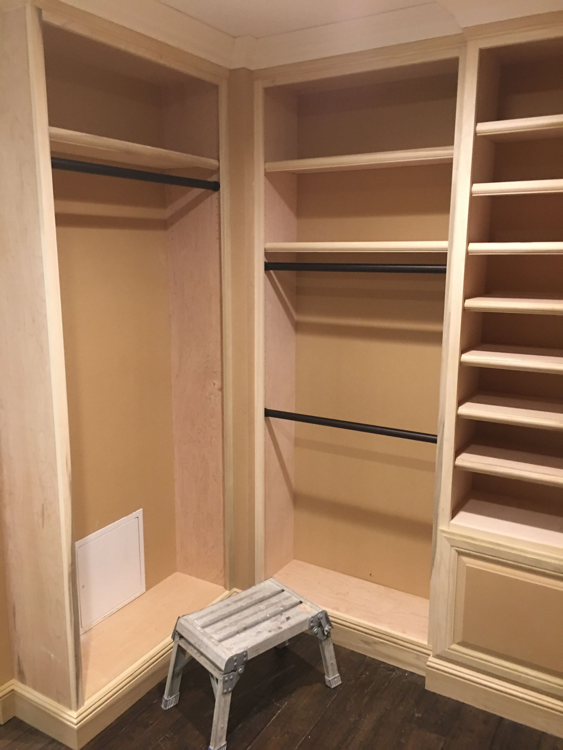 Built-in closet with shelves and hanging rods; small stool in front. Light-colored wood and walls.