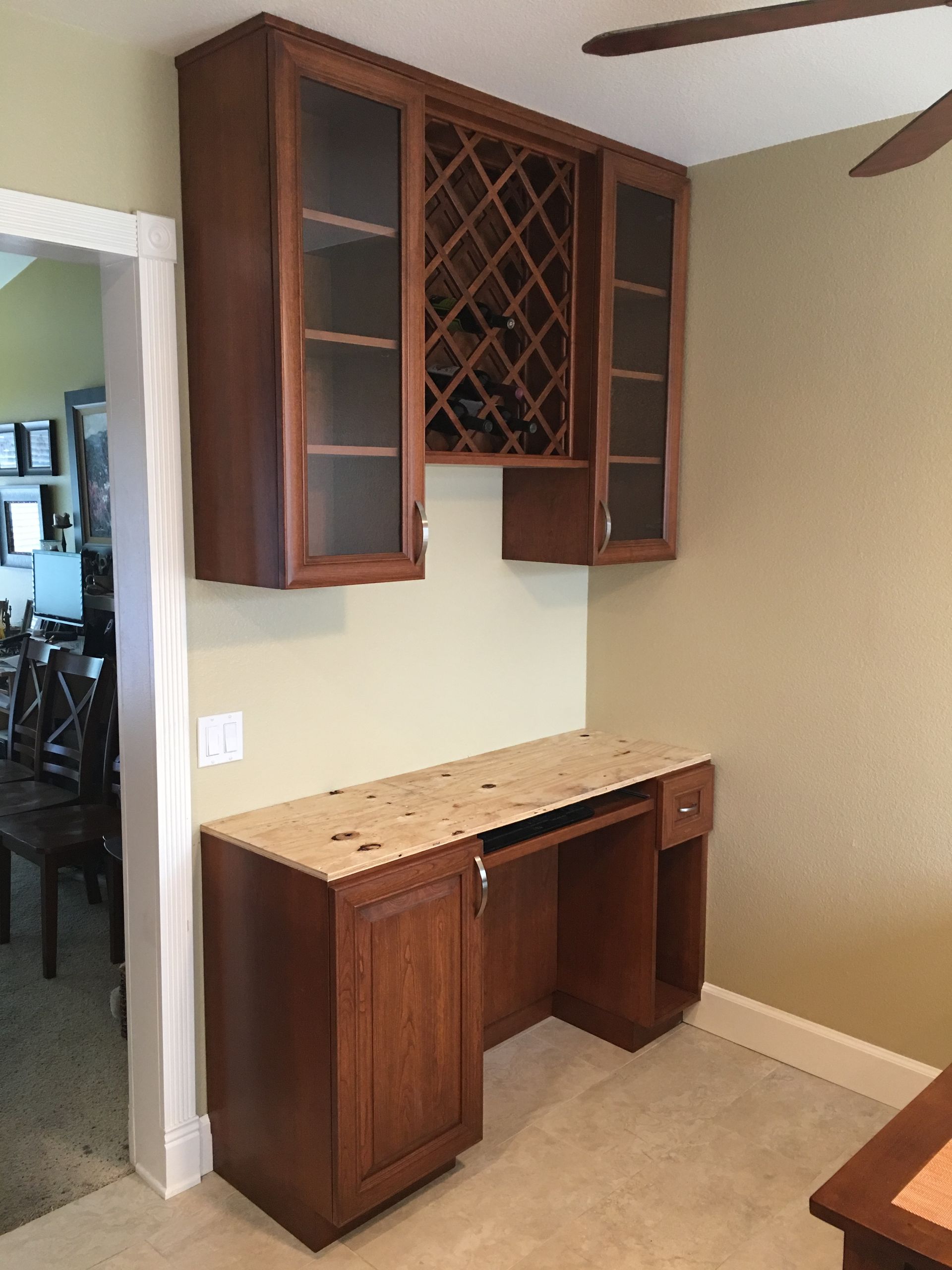 Built-in desk with overhead cabinets: wine rack between glass-front storage, against a beige wall.