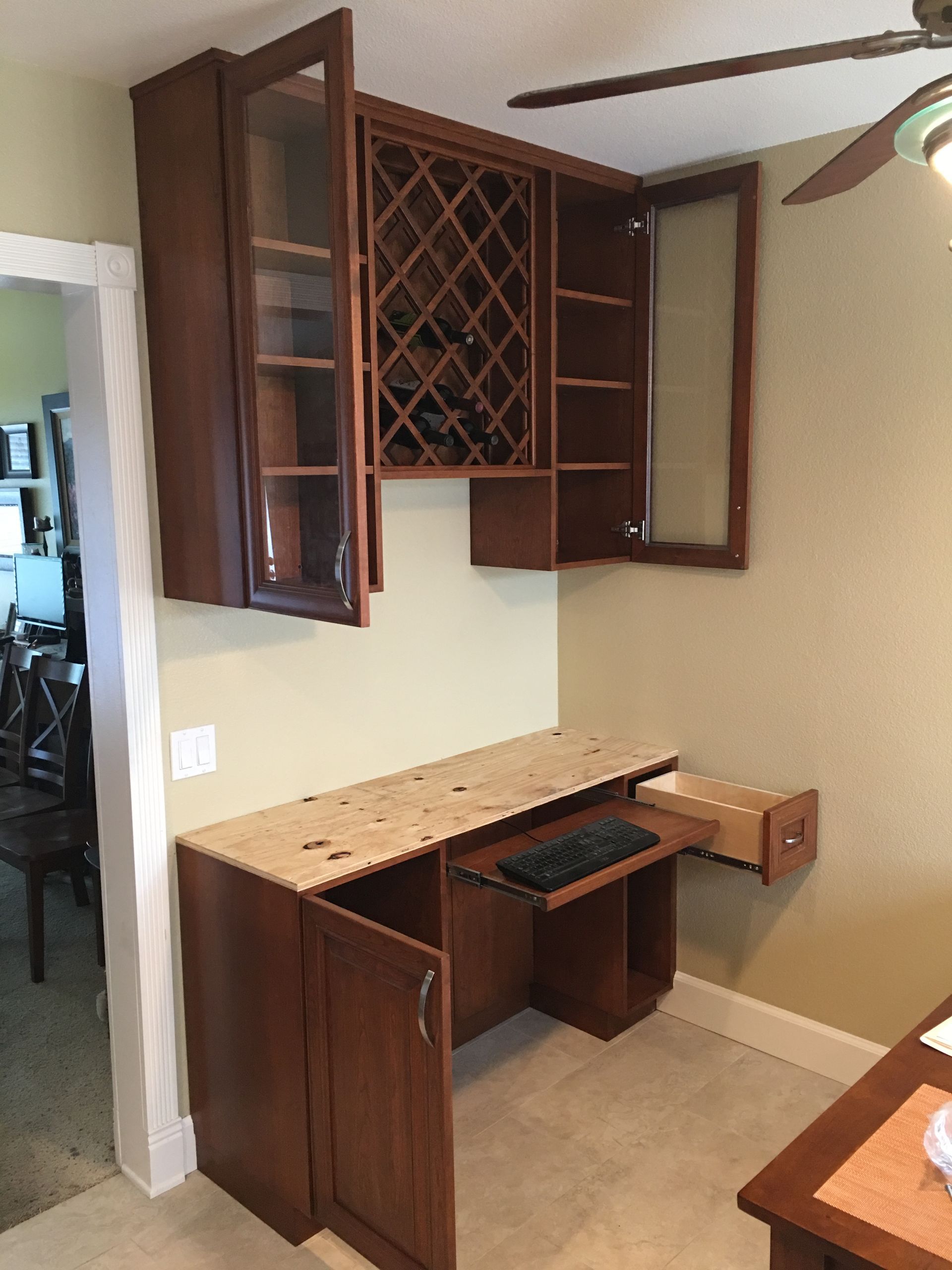 Built-in desk with wine storage, dark brown cabinets, beige countertop, keyboard tray, and glass-door cabinets.