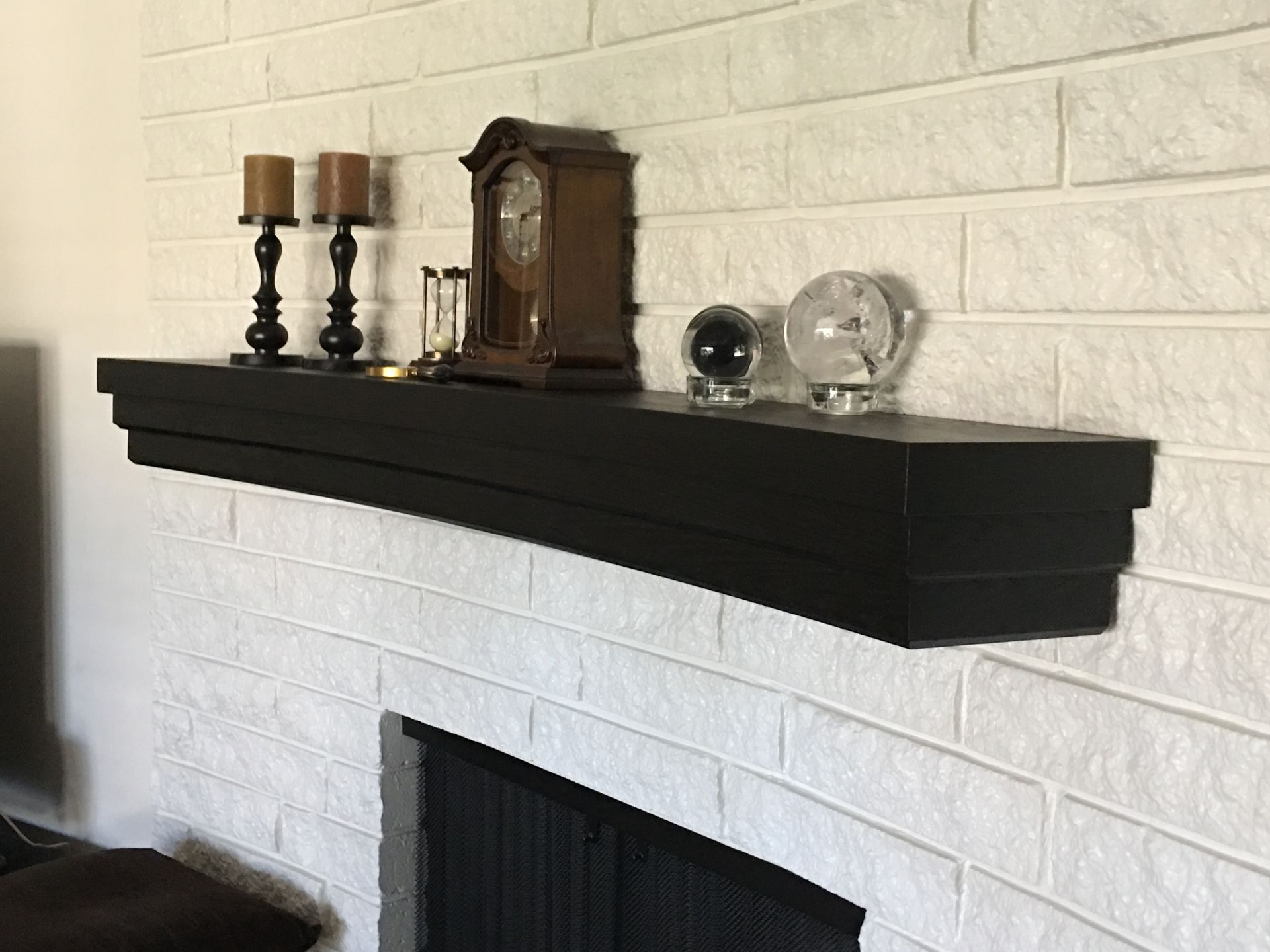 Black shelf with candles, a clock, and decorative spheres, mounted on a white brick wall above a fireplace.