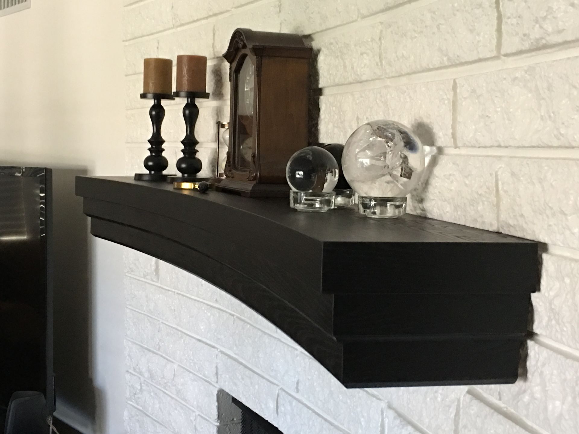 Black fireplace mantel with candles, a clock, and glass orbs, on a white brick wall.