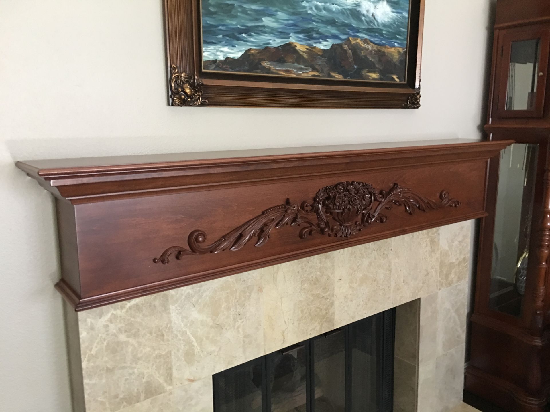 A carved dark wood fireplace mantel over a stone fireplace, with a painting and a grandfather clock nearby.