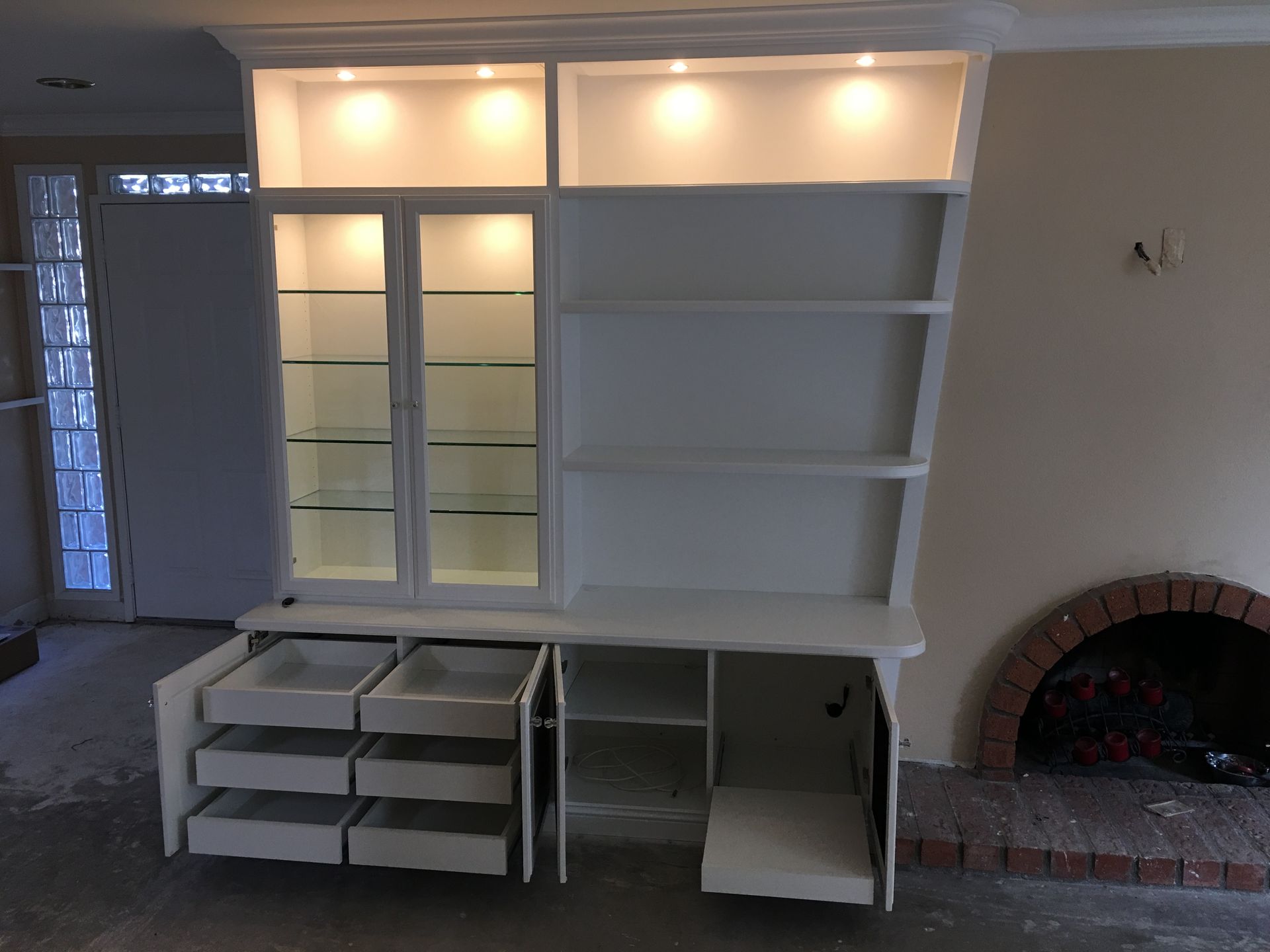 White built-in cabinet with glass-doored display, shelves, and drawers, next to a fireplace.