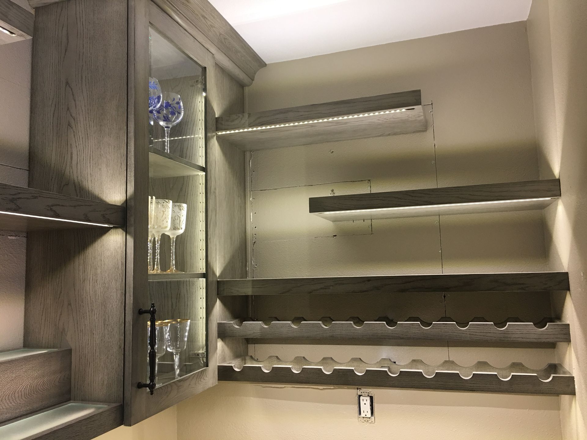 Gray built-in shelving and cabinet with LED lighting; wine rack and glass display.