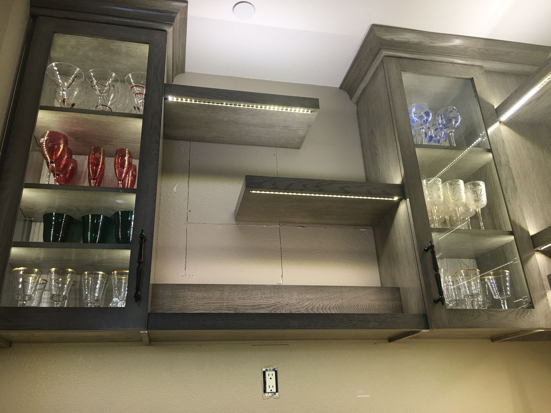 Kitchen cabinet with glass shelves, lit with LED lights, displaying glassware. Gray cabinets, neutral wall.
