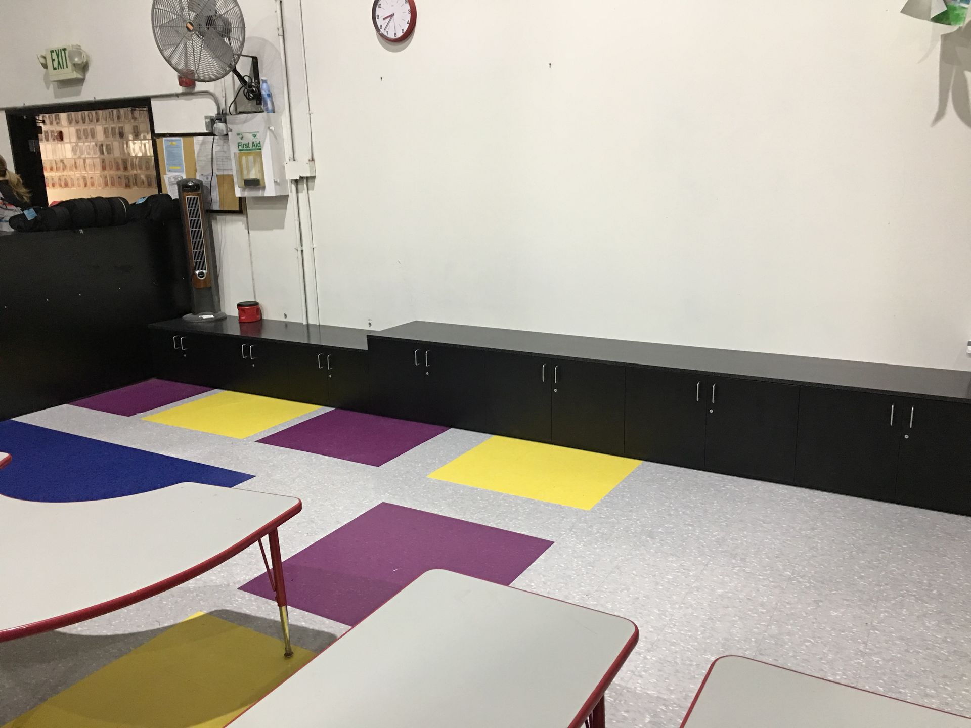 Classroom with low black cabinets along a white wall, colorful floor tiles, and child-sized tables.