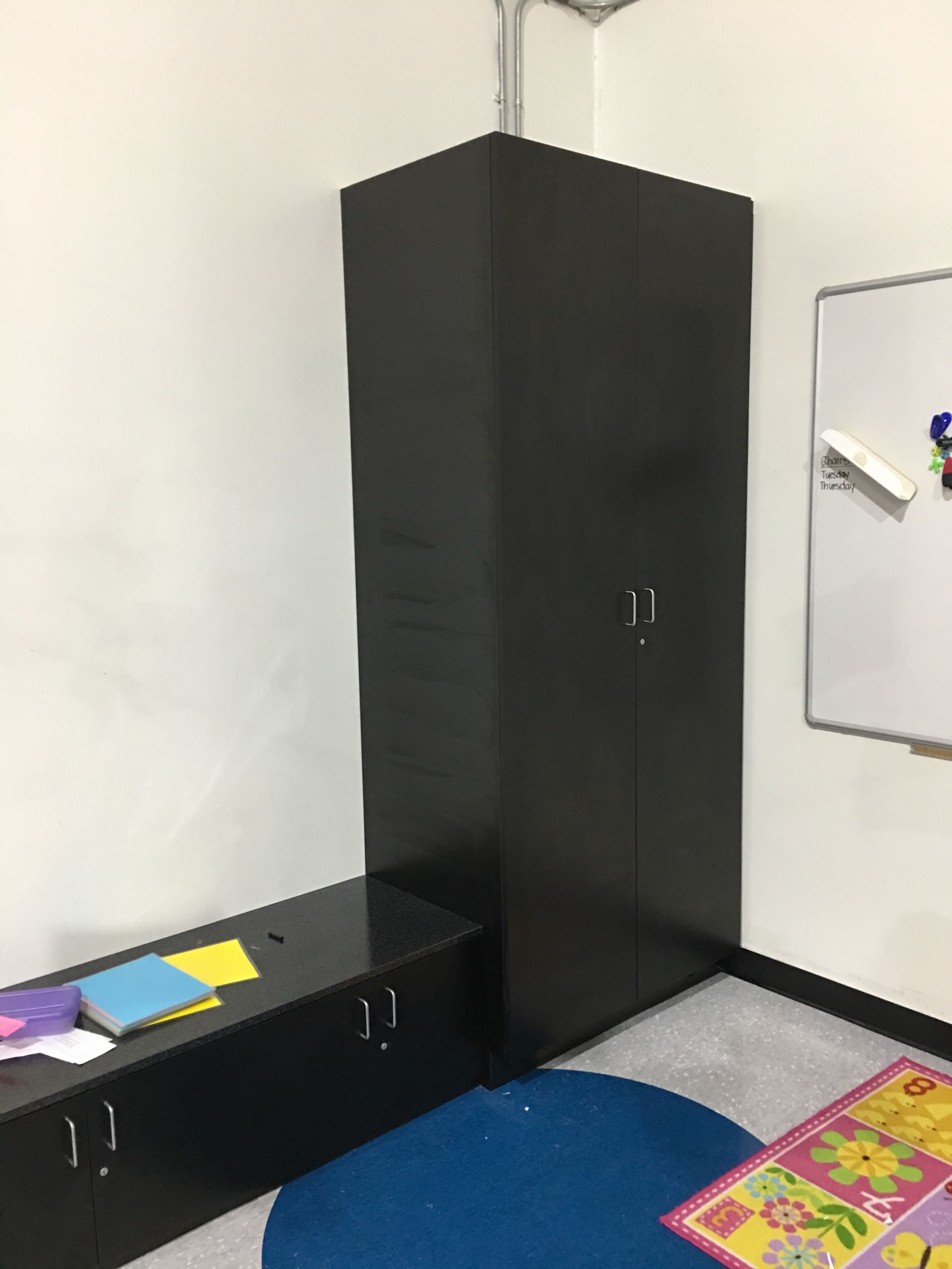 Black storage cabinets and bench in a corner of a room, next to a whiteboard and a blue circle rug.