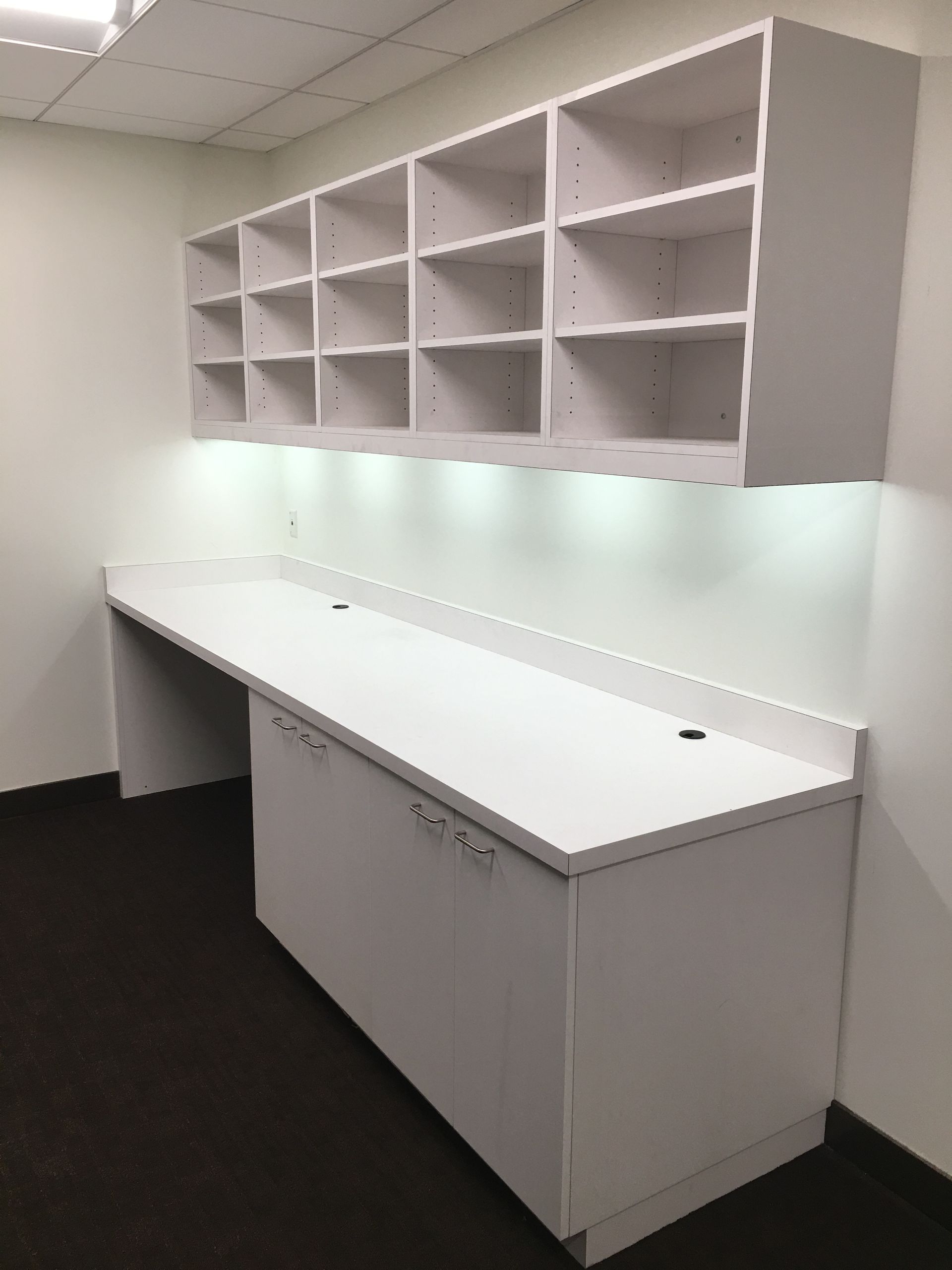 White built-in desk with overhead shelves and under-shelf lighting in a room with dark carpet and white walls.