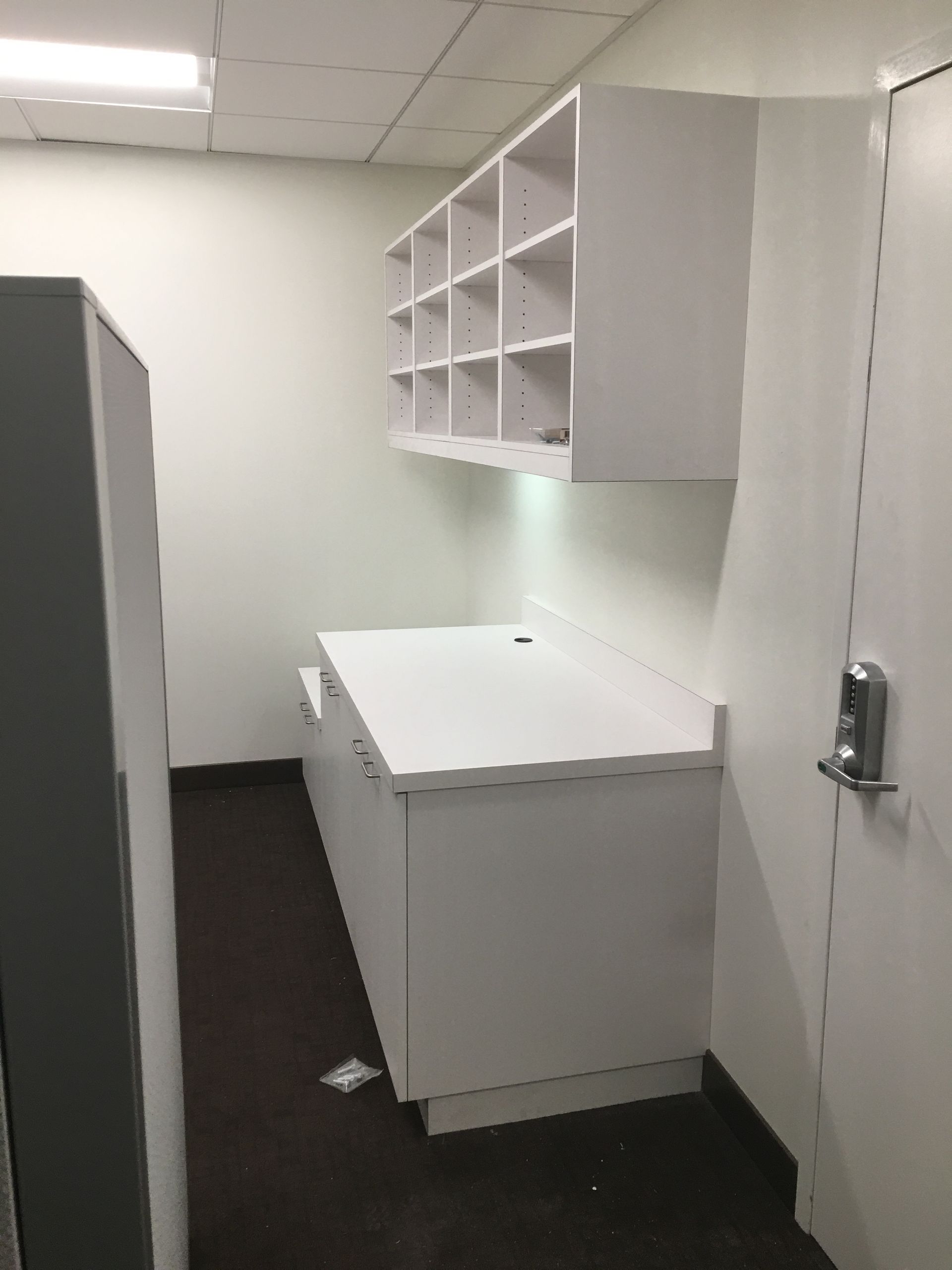 White cabinets and shelving unit in a narrow room with a dark floor, next to a white door and gray object.
