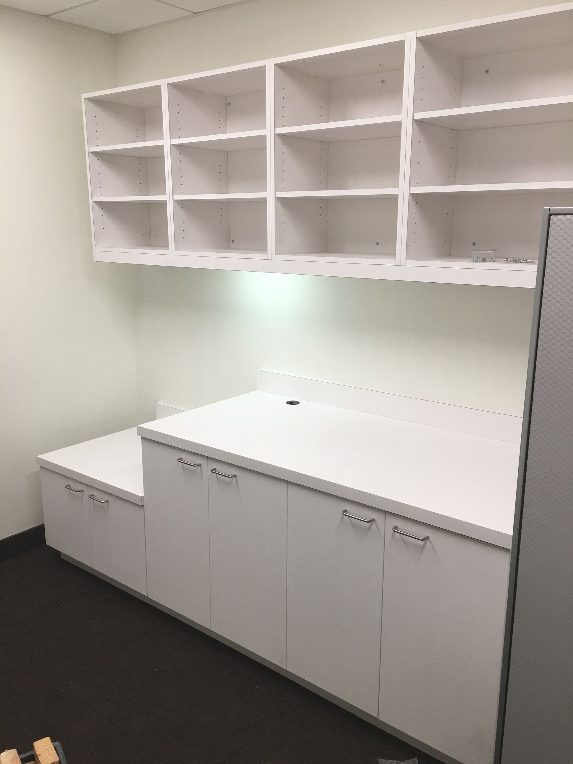 White built-in office furniture, including a desk, cabinets, and overhead shelving, against a white wall.