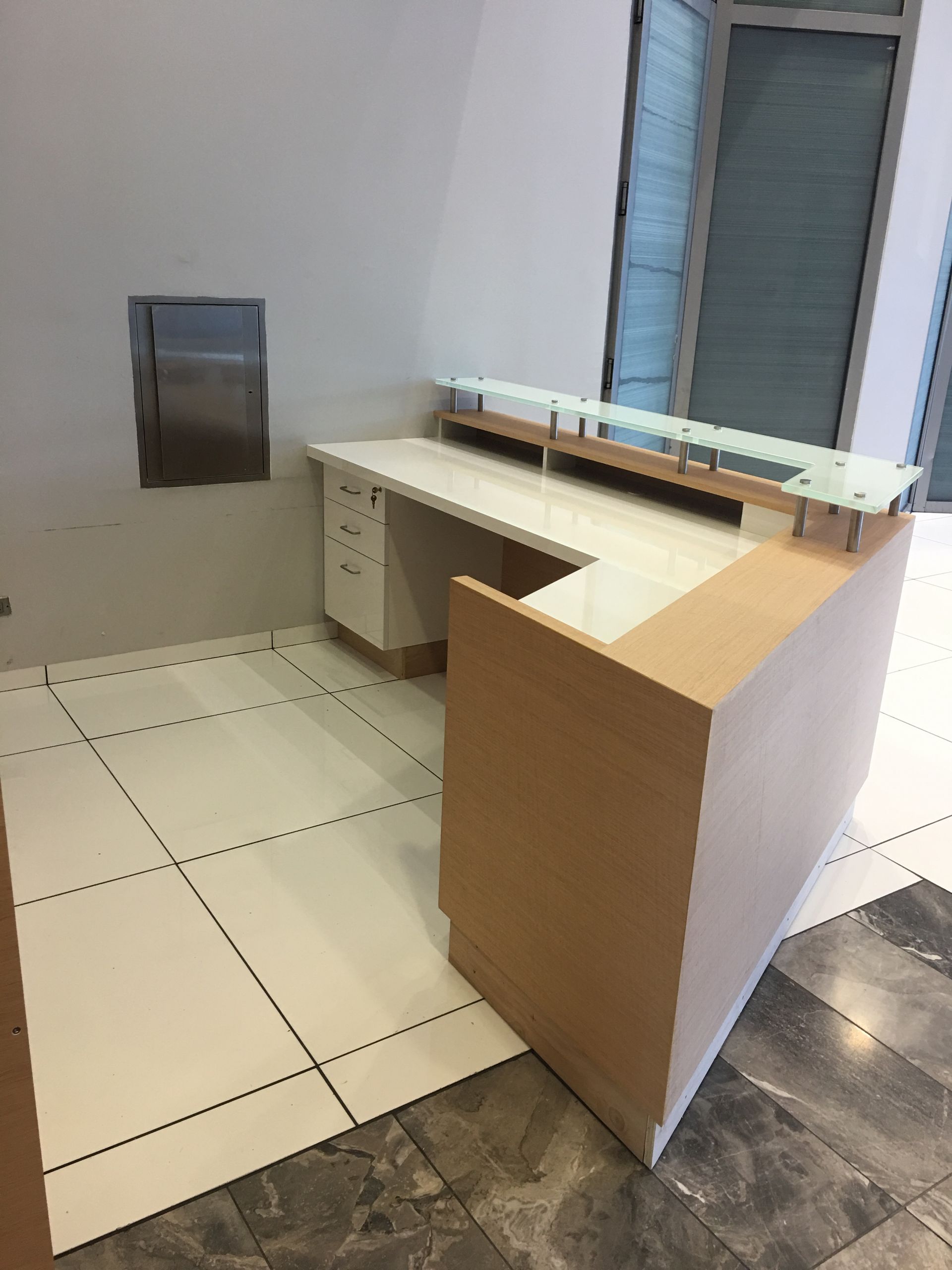 Reception desk in a corner, light wood and white surface, glass top. Beige and white tiled floor, window in the background.