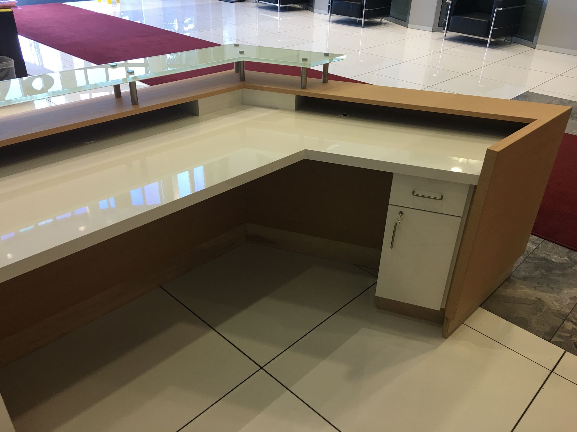 Beige and wood L-shaped reception desk with a glass shelf, in a tiled room.