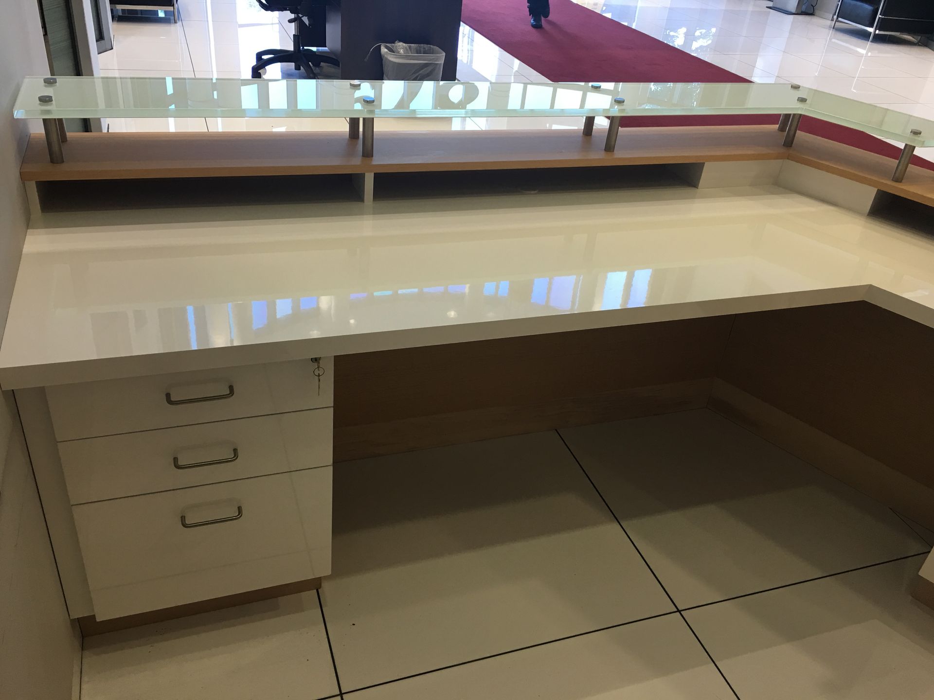 White reception desk with three drawers, glass panel, and built-in shelving in a well-lit space.