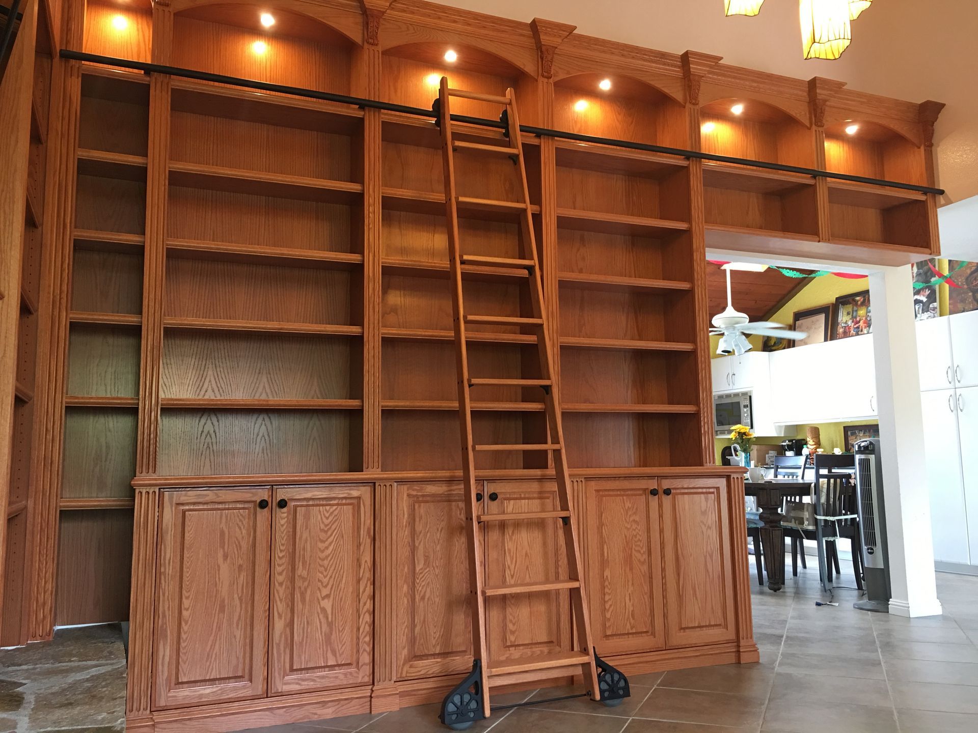 Built-in wooden bookshelves with rolling ladder, lit by spotlights. Cabinets on lower level. Dining room in background.