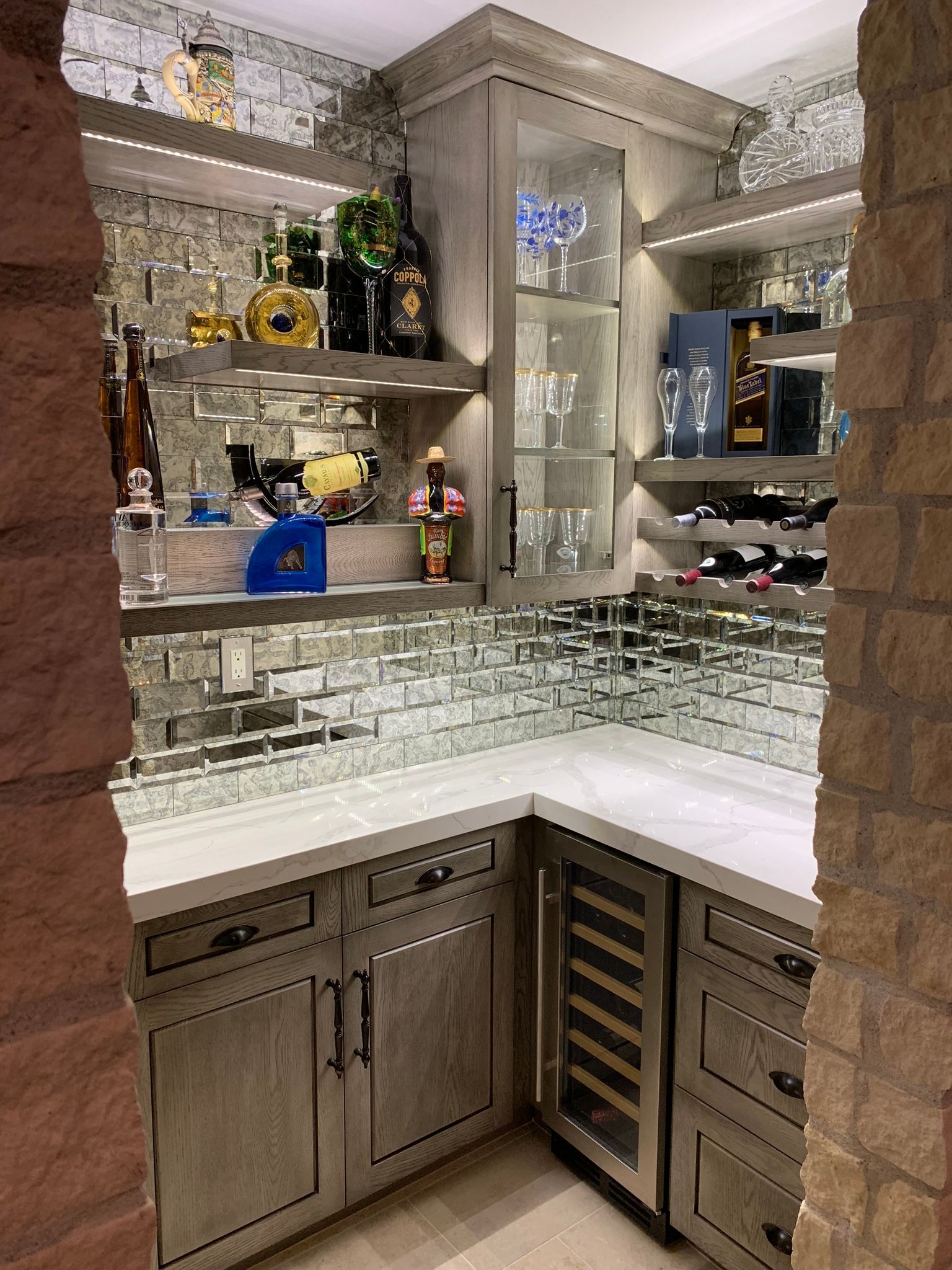 Corner home bar with mirrored walls, shelves stocked with bottles, and a built-in wine fridge.