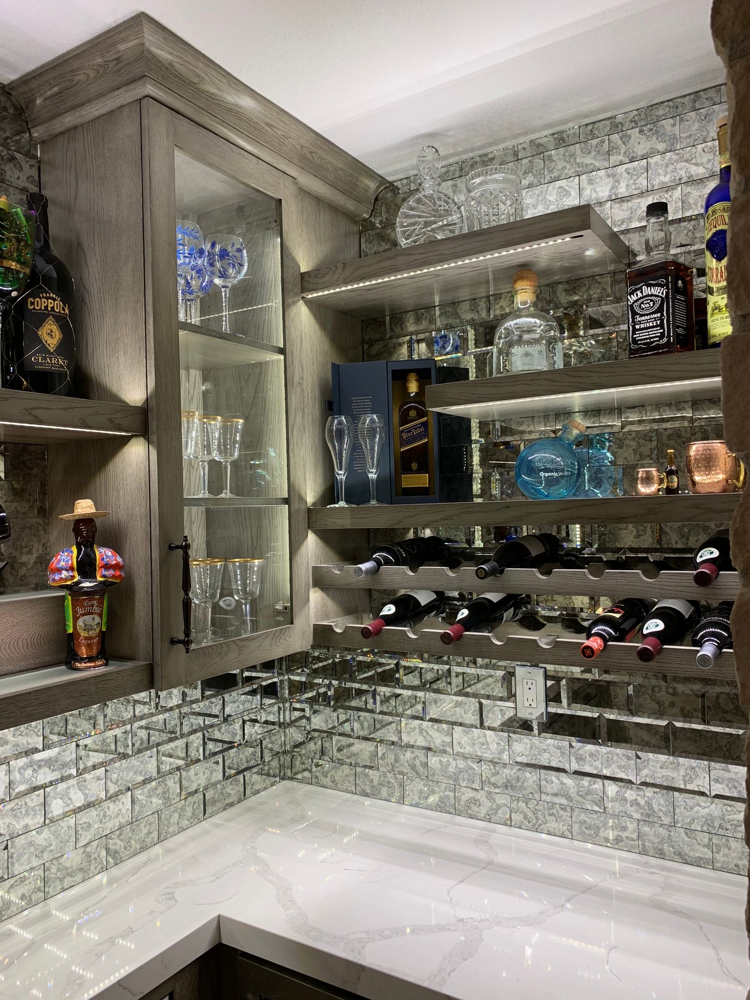 Well-stocked, lit home bar with liquor, glassware, and wine racks set against a mirrored brick wall.